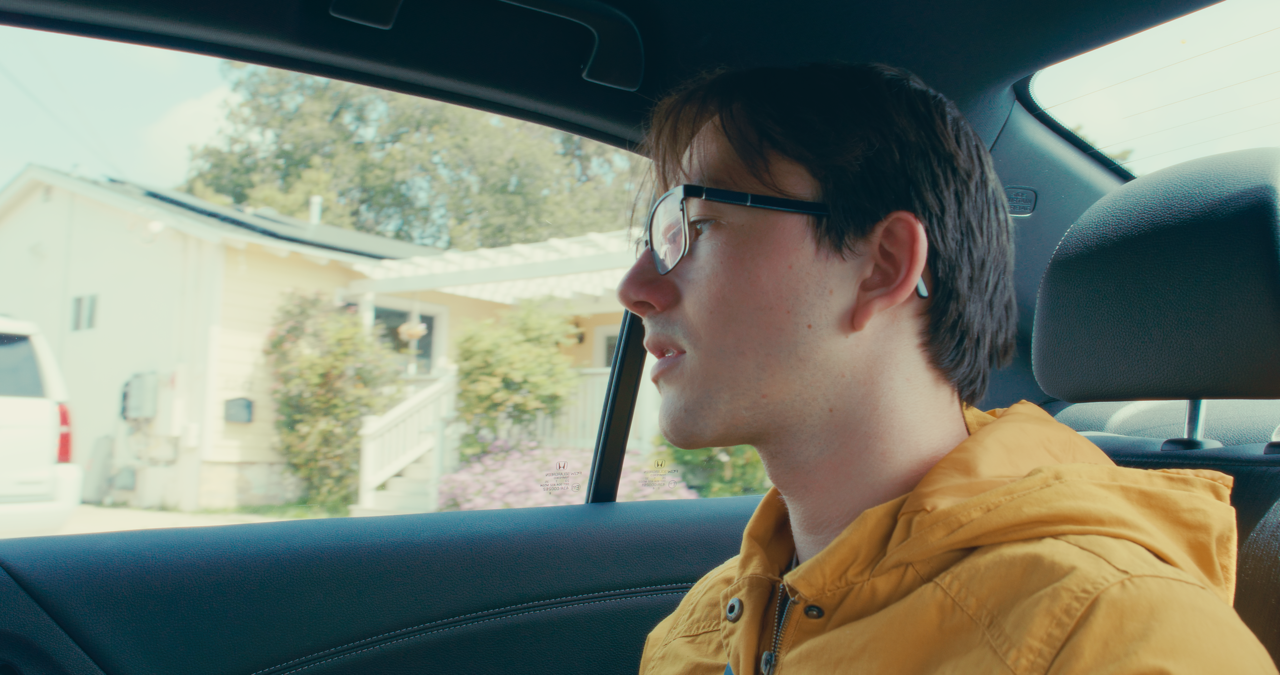 Young man with dark hair, glasses, and wireless earbuds sitting in the backseat of a car, wearing a yellow jacket.