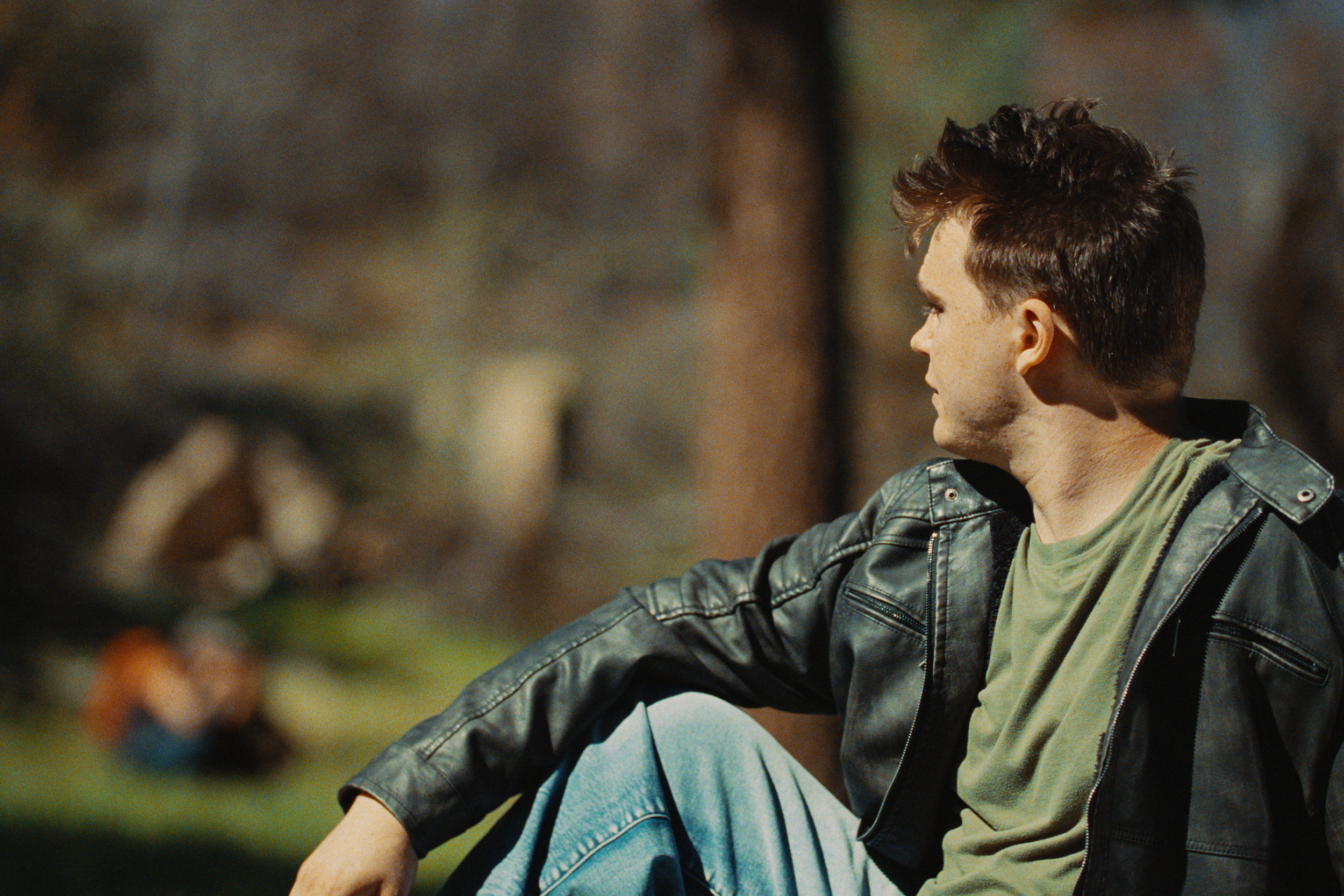 Profile of a young man with dark, curly hair wearing a black leather jacket and a green t-shirt, sitting outside.