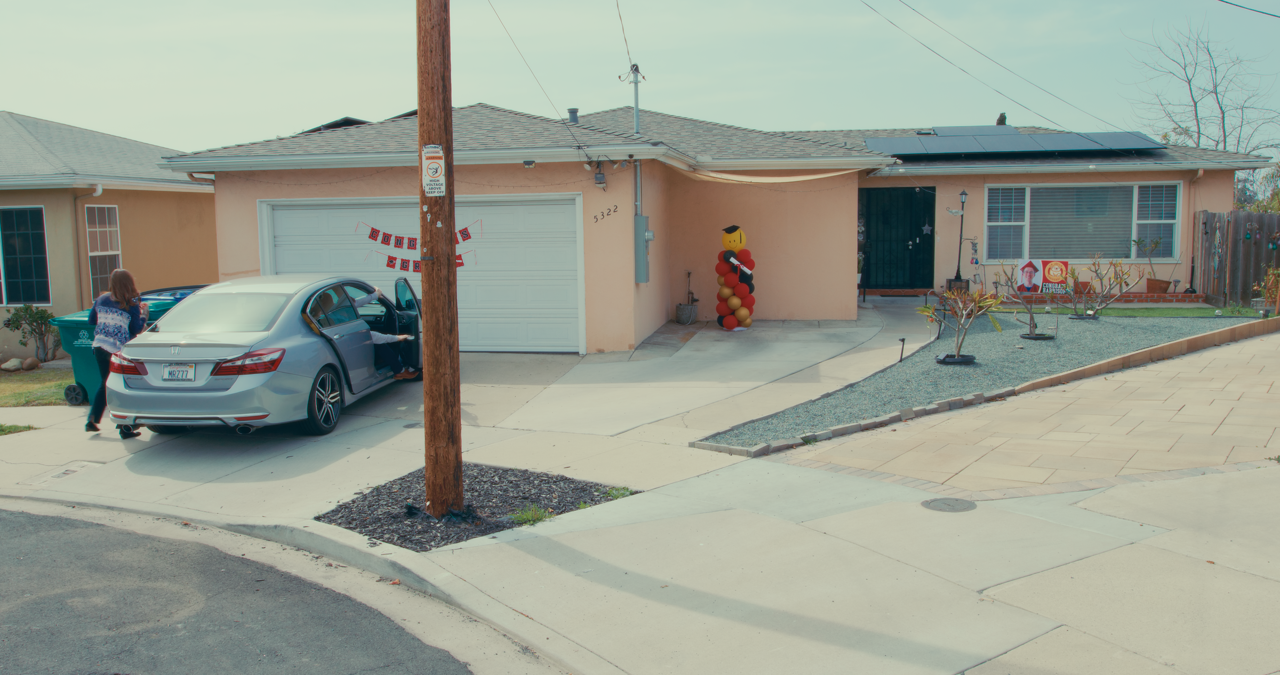 A person putting groceries into the trunk of a silver car parked in a driveway in front of a peach-colored house with a decorated balloon figure near the entrance.