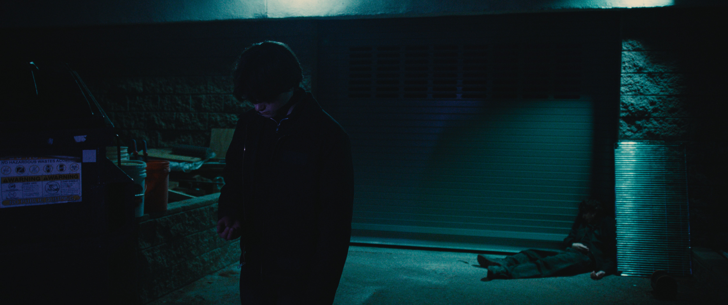 A person standing next to a waste disposal area with a homeless person lying on the ground nearby at night.