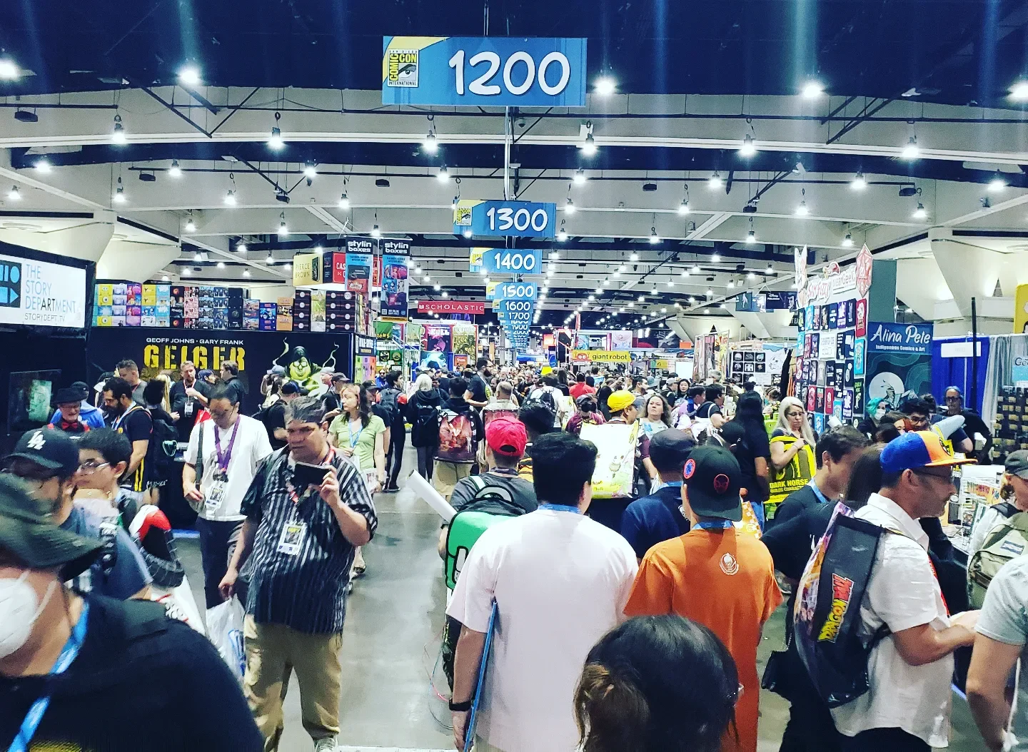 Crowd of people at a comic convention inside a large hall with booths, banners, and signs indicating seating sections 1200, 1300, 1400, and 1500, and displays featuring various merchandise and artwork.