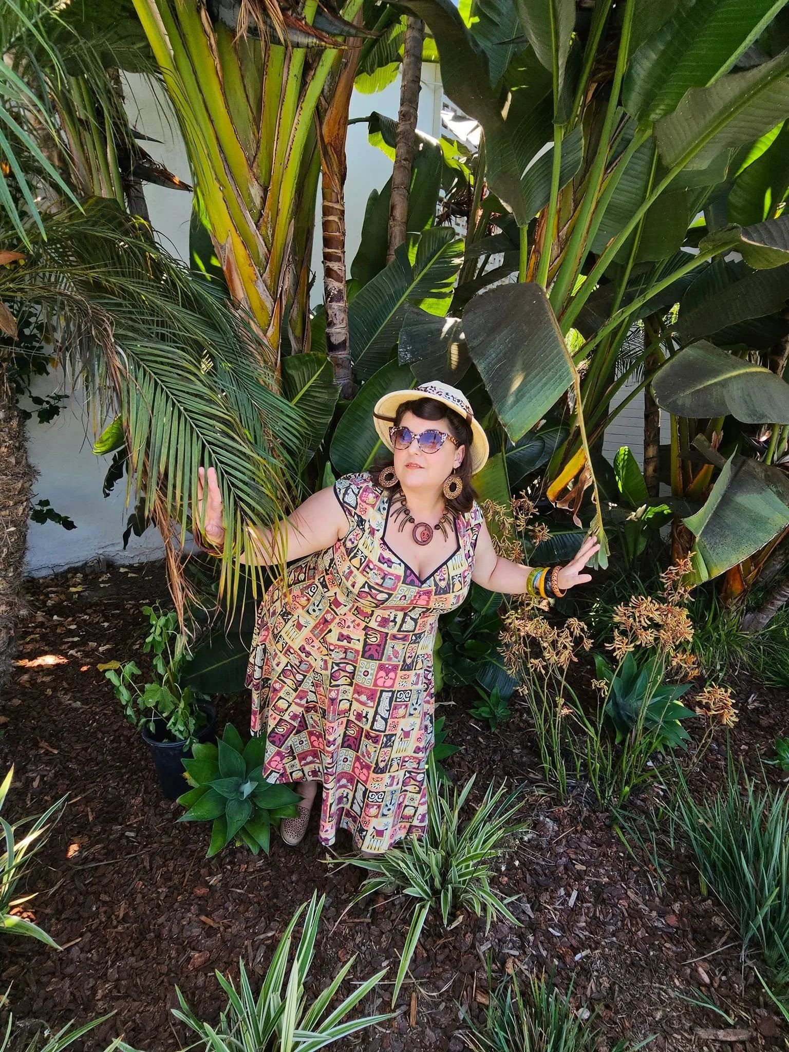 A woman in a colorful patterned dress, wearing sunglasses, a hat, and various jewelry, standing among tropical green plants in a garden.