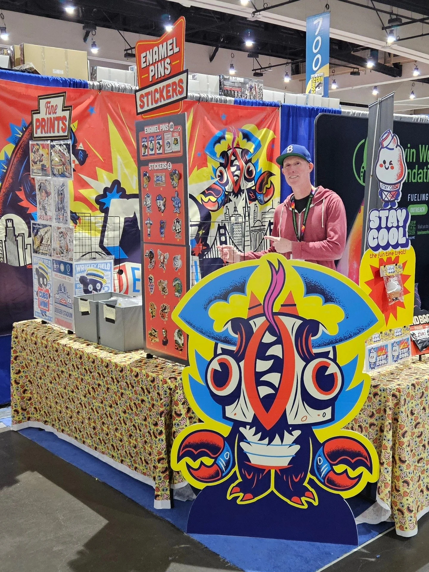 A convention booth featuring colorful artwork, enamel pins, stickers, and prints related to a Lovecraftian theme, with a person smiling and pointing at the display.
