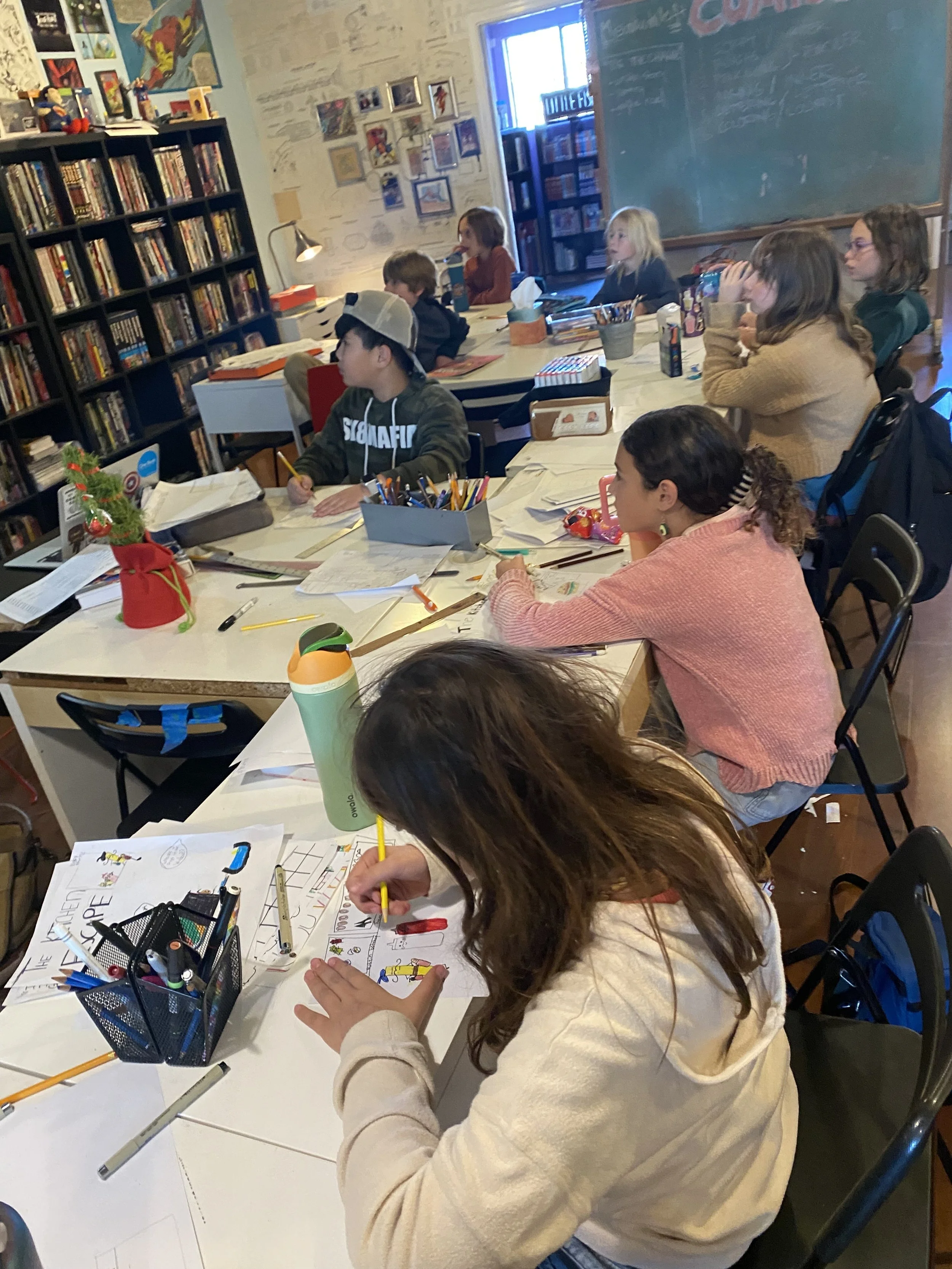 Children sitting at a table in a classroom, working on drawings and projects with various supplies, walls decorated with artwork and bookshelves in the background.