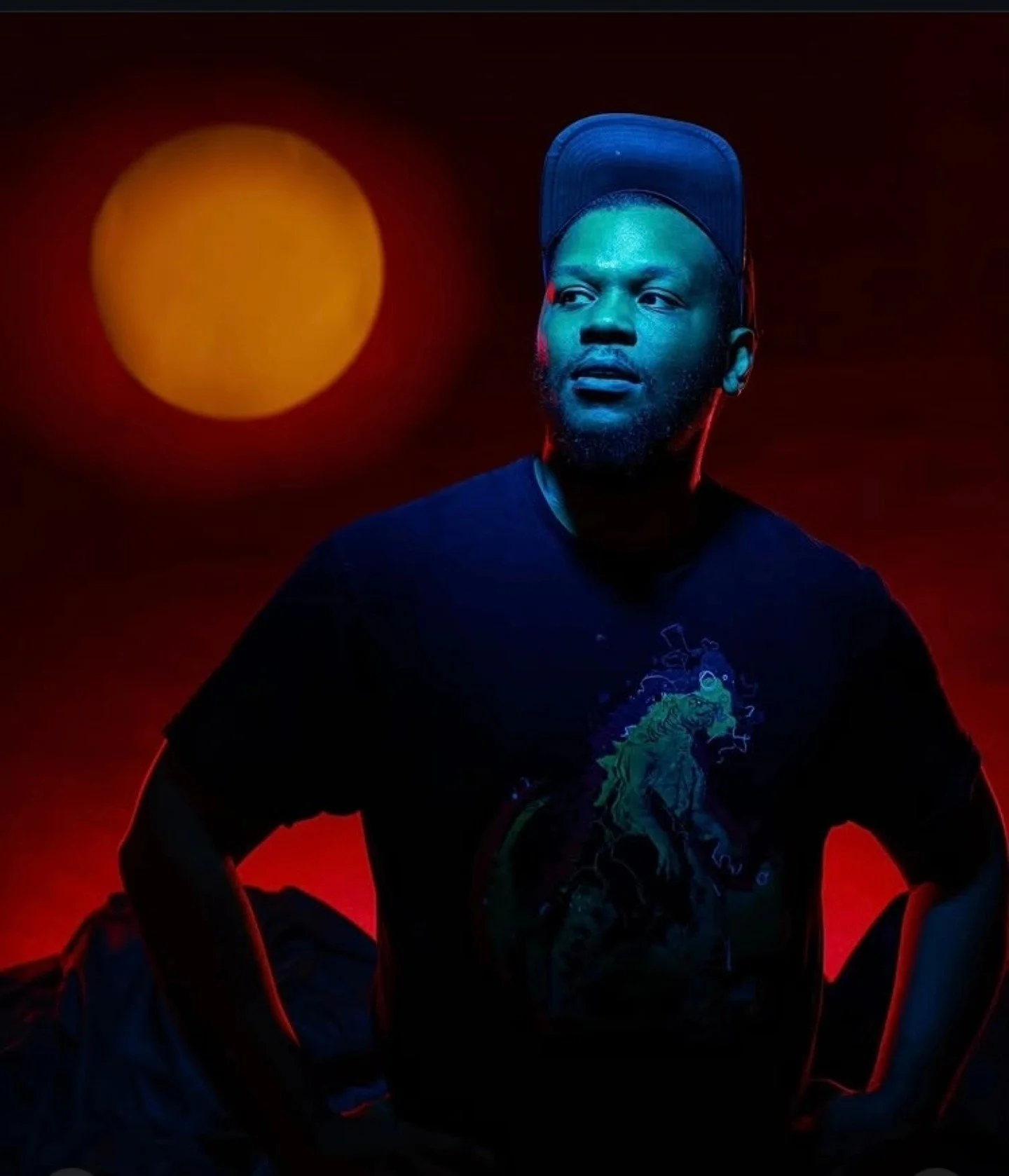A young man with a beard and a baseball cap worn backwards, illuminated by blue and red lighting, sits cross-legged with hands on his knees, against a dark background featuring a large orange circle resembling a sun or moon.