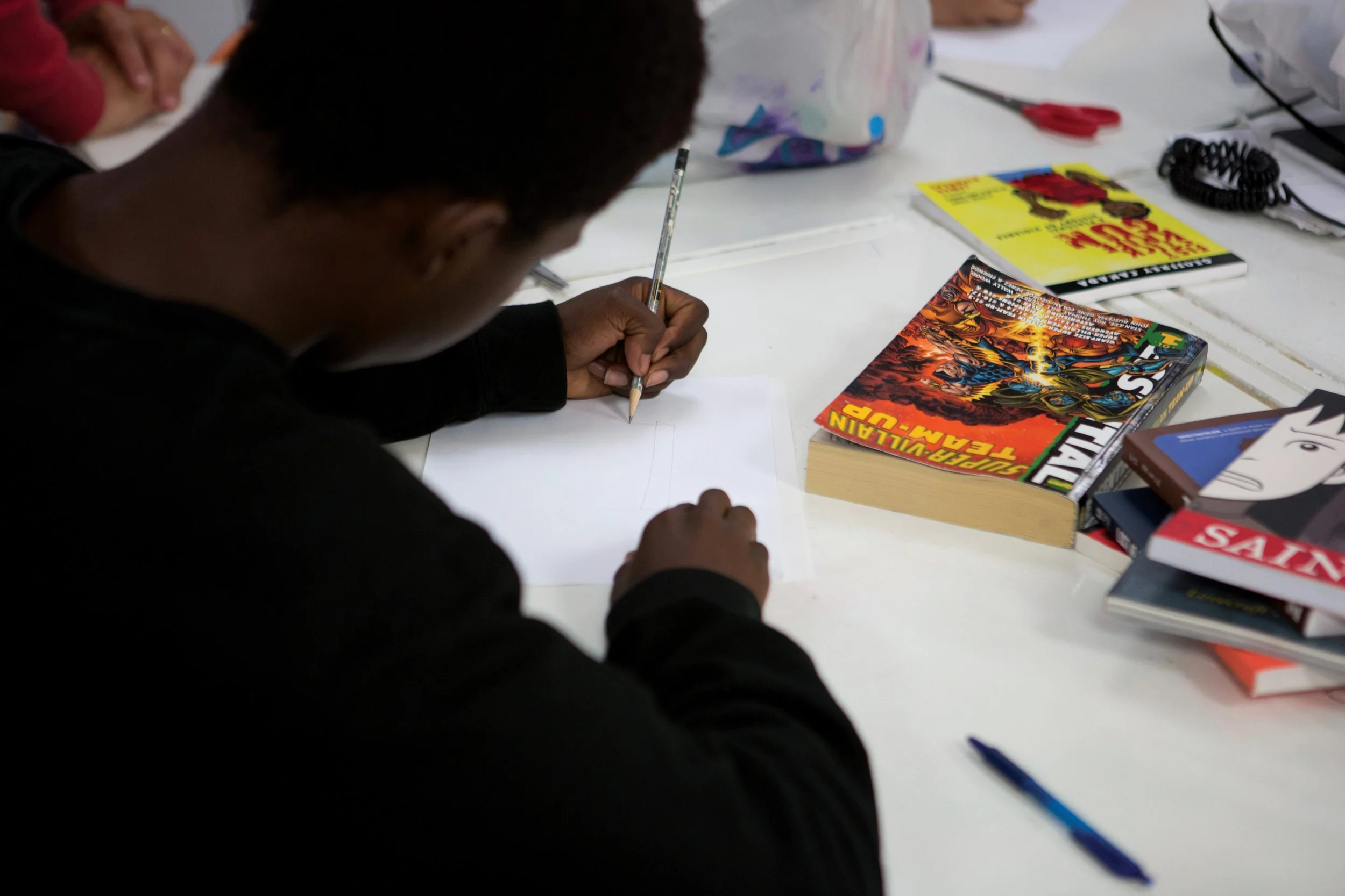 A person with dark hair sitting at a table, writing on a piece of paper with a pencil. The table has various books, including one titled "Supervillain Team-Up" and others related to comic books. There are also some pens, scissors, and a black coiled cord on the table.