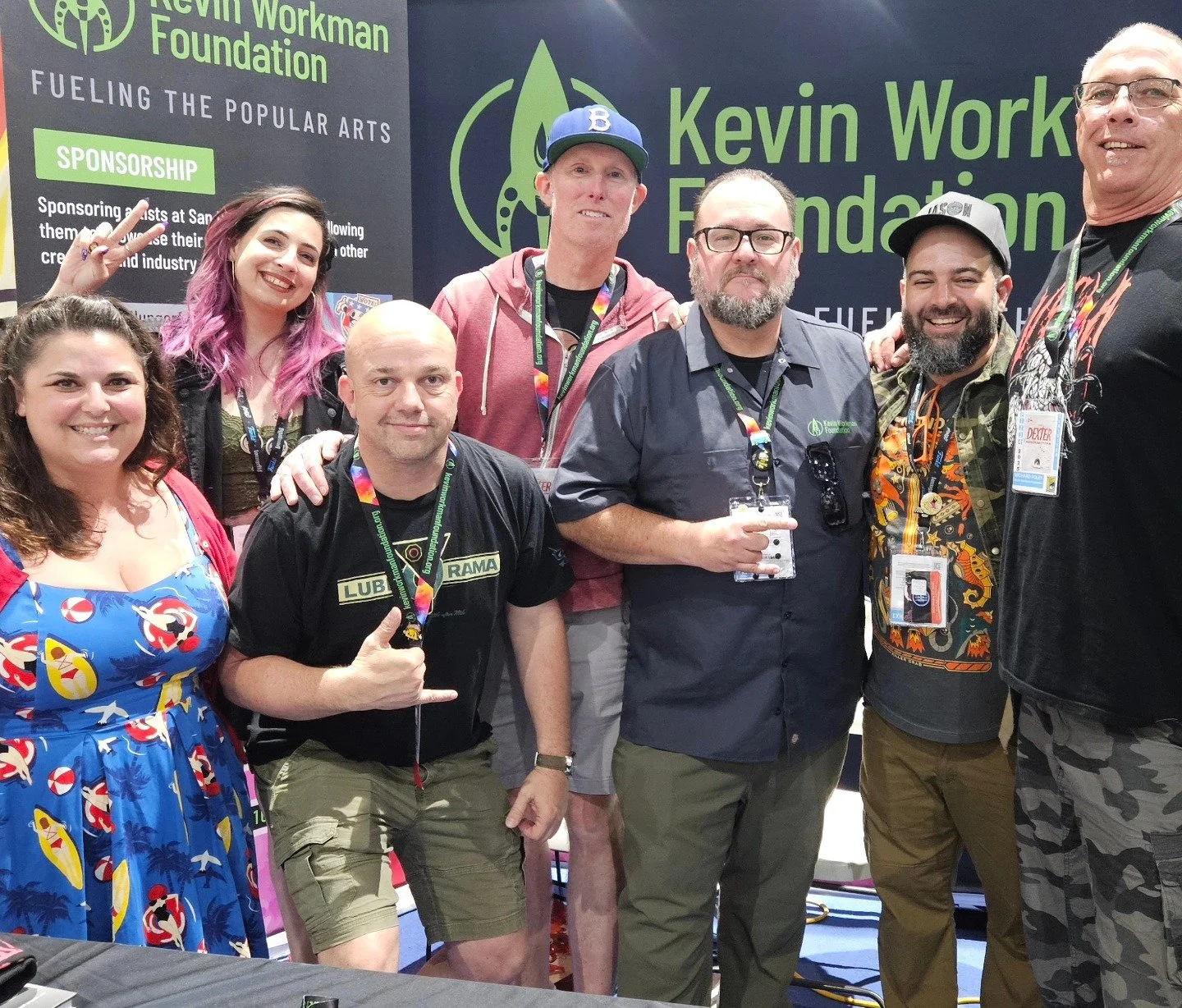 Group of eight people posing together at an event, in front of a backdrop with the logo and name of the Kevin Workman Foundation. They are smiling and wearing various casual outfits, some with event badges and lanyards.