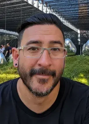 A man with dark hair, glasses, and a beard smiling outdoors near metallic sculptures.