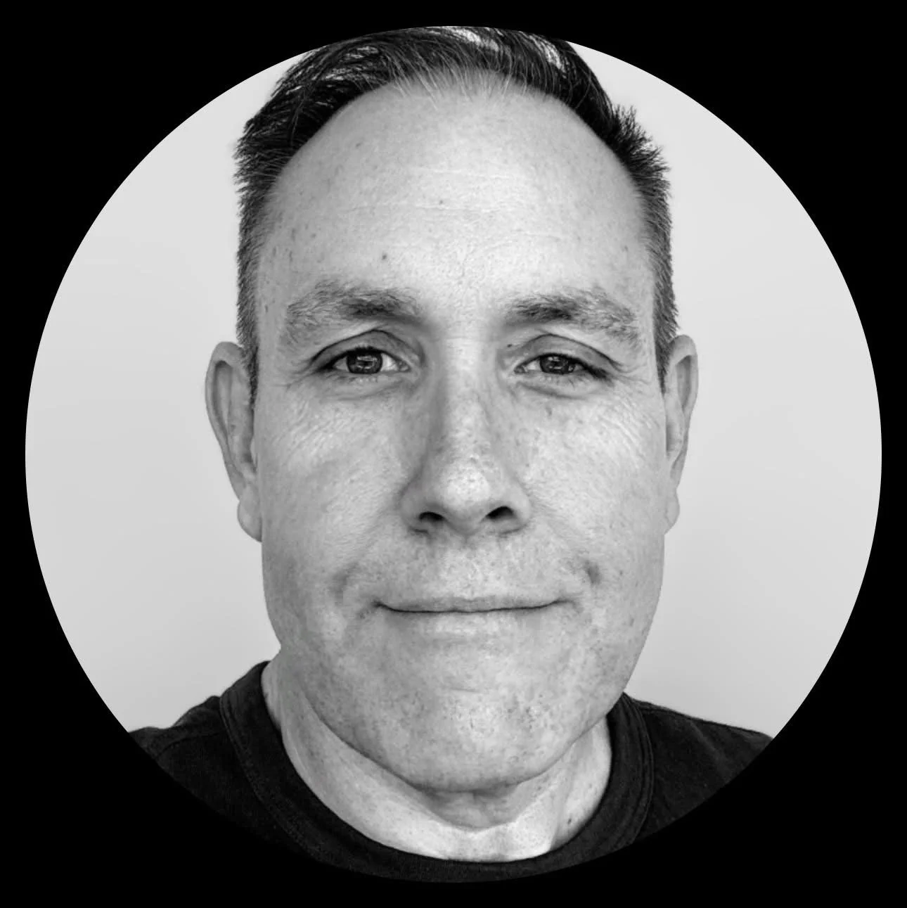 Black and white photo of a smiling man with short dark hair, wearing a black shirt, facing forward.