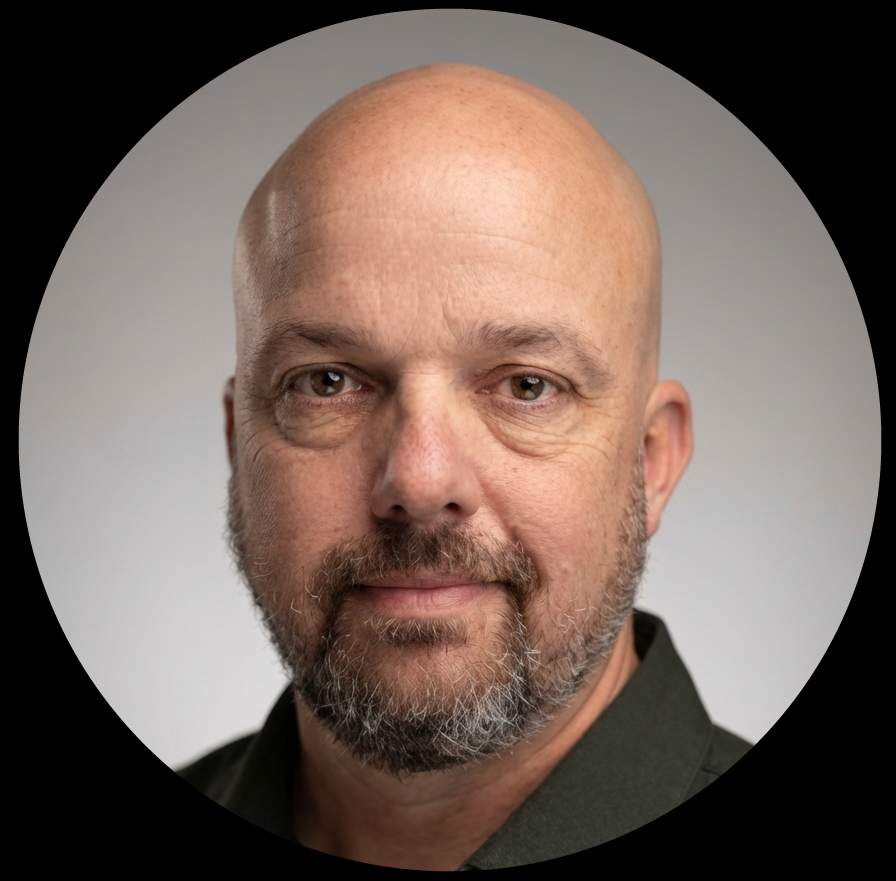 Close-up of a man with a bald head and a beard, wearing a dark shirt, against a neutral gray background.