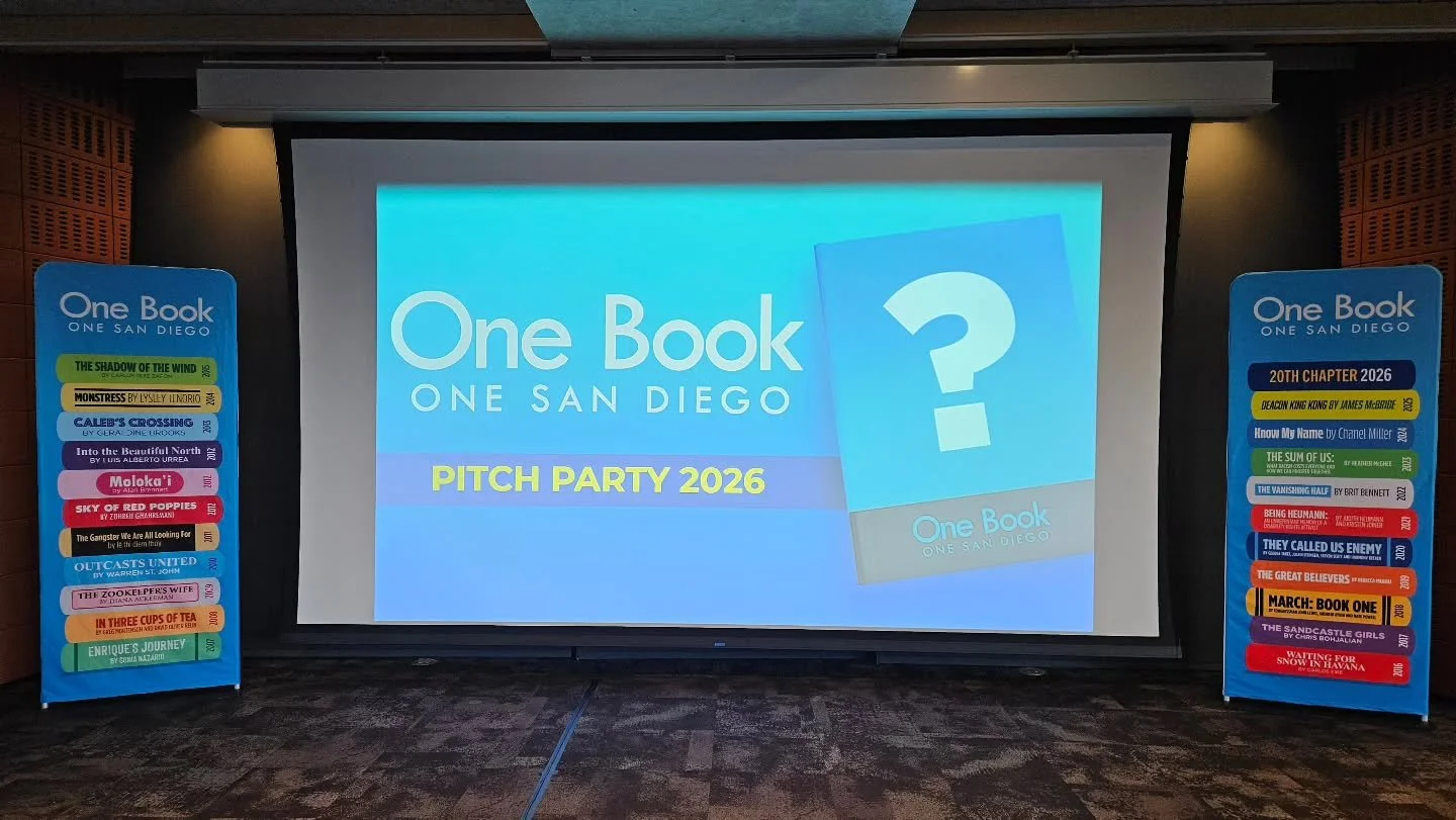 A presentation screen displays the text 'One Book One San Diego PITCH PARTY 2026' in a conference room flanked by two vertical banners listing book titles.