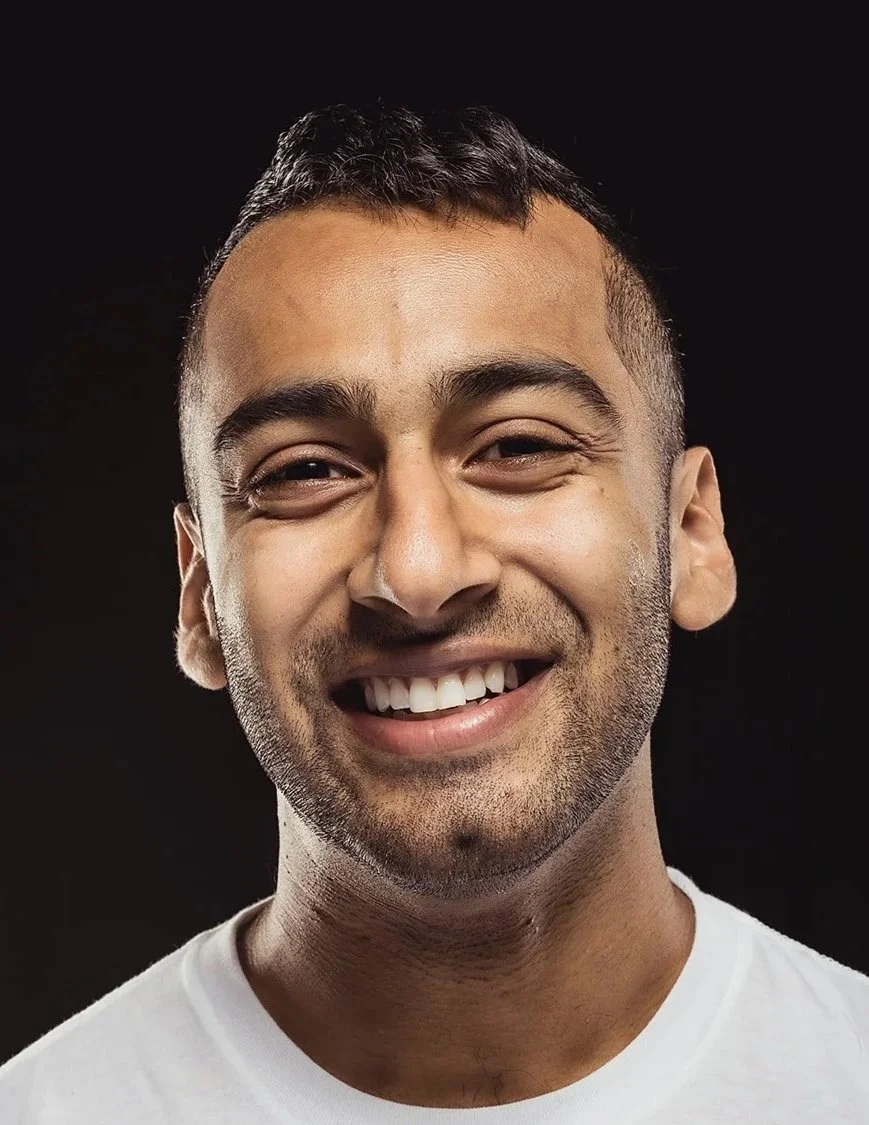 Close-up of a smiling young man with short dark hair and stubble, wearing a white shirt against a black background.