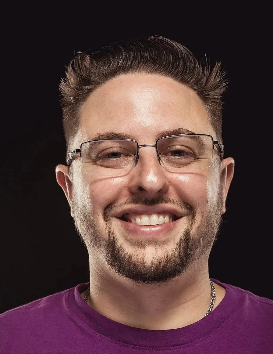 A smiling man with glasses, facial hair, and a purple shirt standing against a black background.