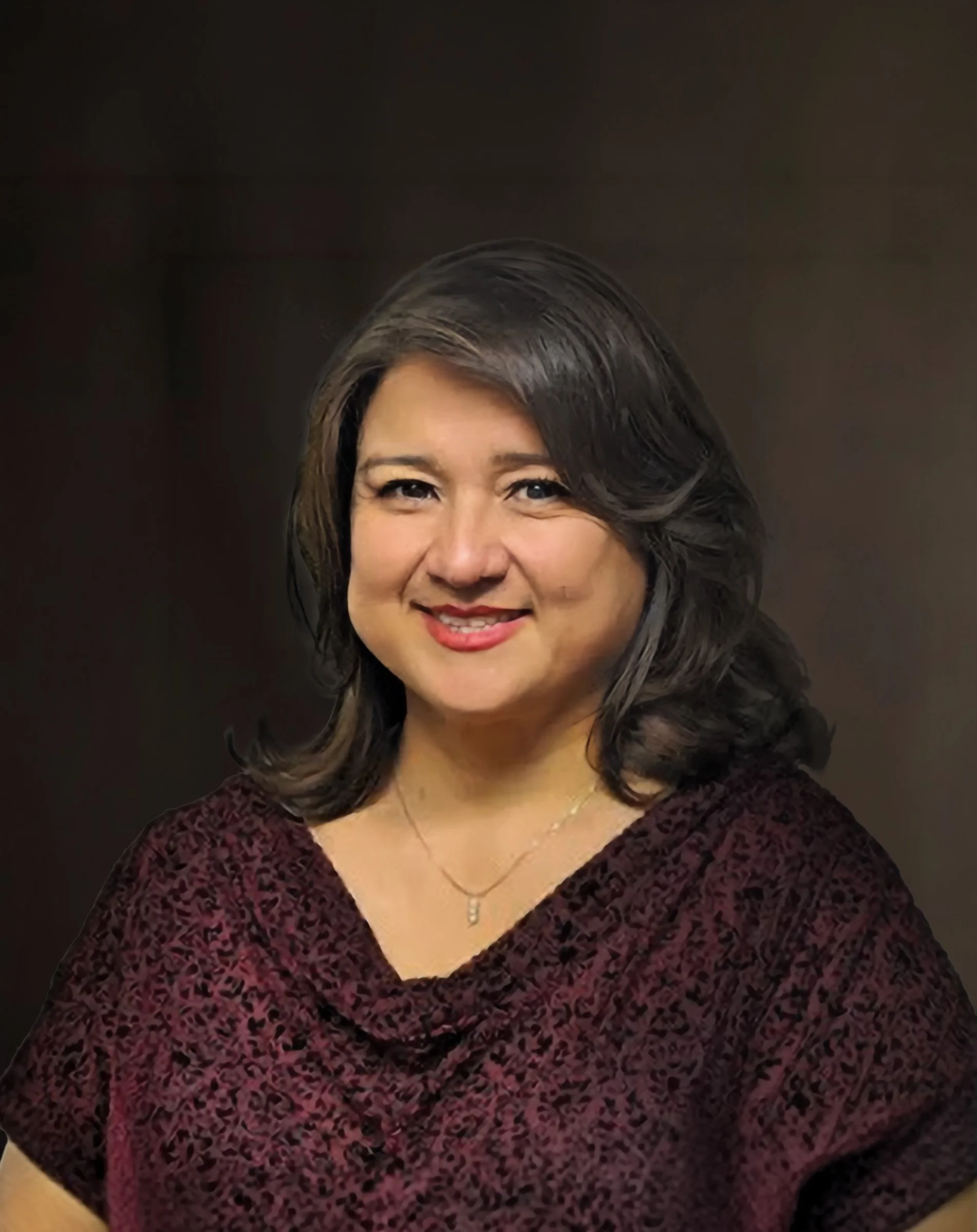 Portrait of a woman with shoulder-length dark hair, smiling, wearing a burgundy patterned top and a delicate necklace.