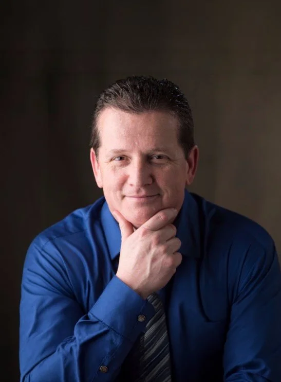 Man in blue shirt with striped tie, posing with hand on chin and smiling lightly.