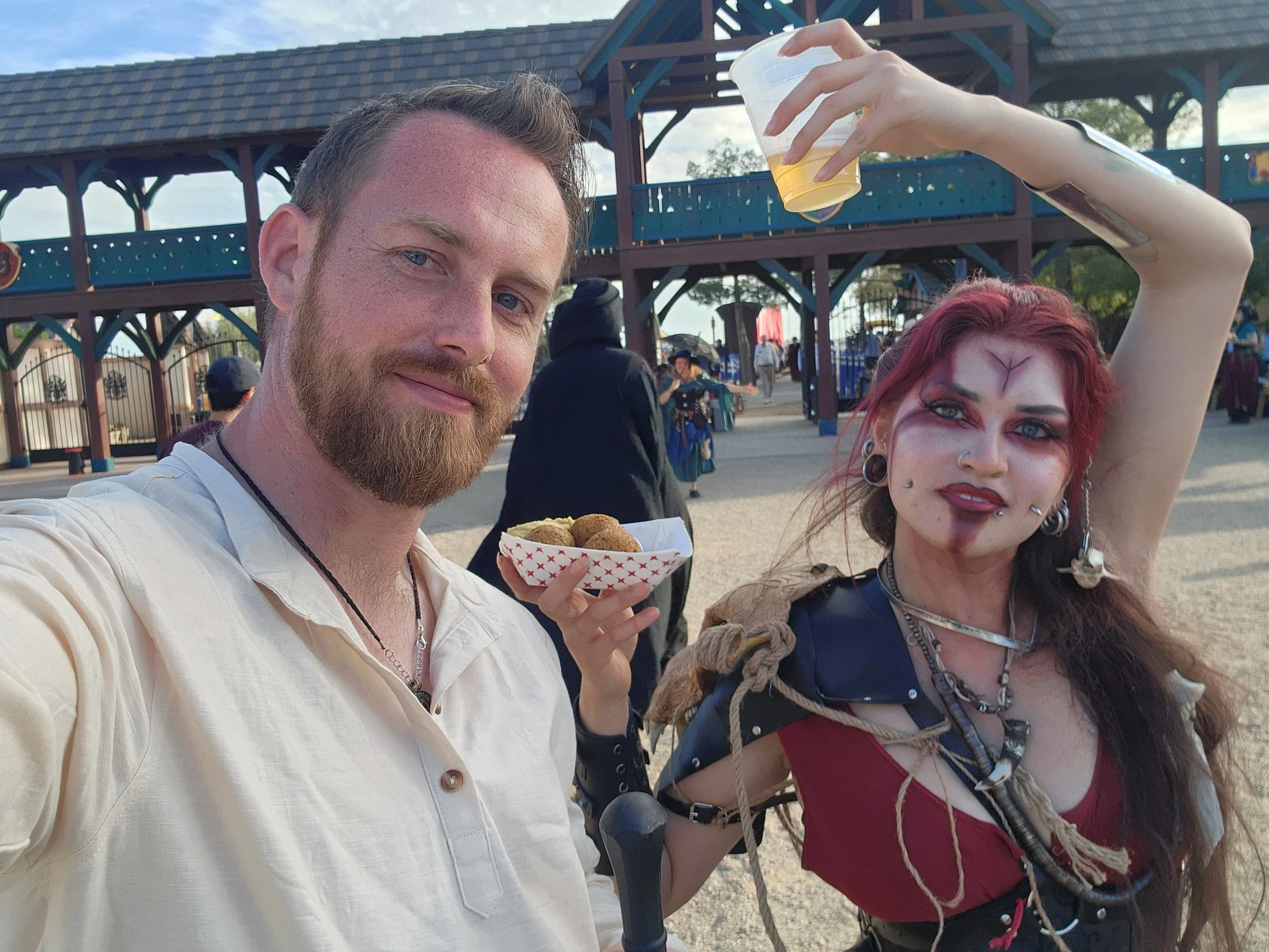 A man and a woman in elaborate costumes taking a selfie at an amusement park. The man is smiling and holding a tray of food, while the woman has dramatic makeup, piercings, and accessories, with her arm raised holding a cup.