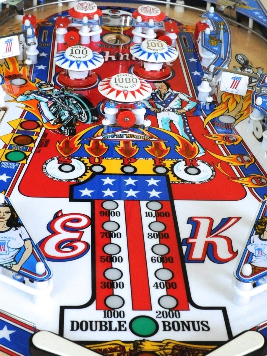 Close-up of a vintage Evel Knievel pinball machine with a patriotic American theme, featuring images of a motorcycle rider, an eagle, and an American flag, with colorful bumpers and targets.