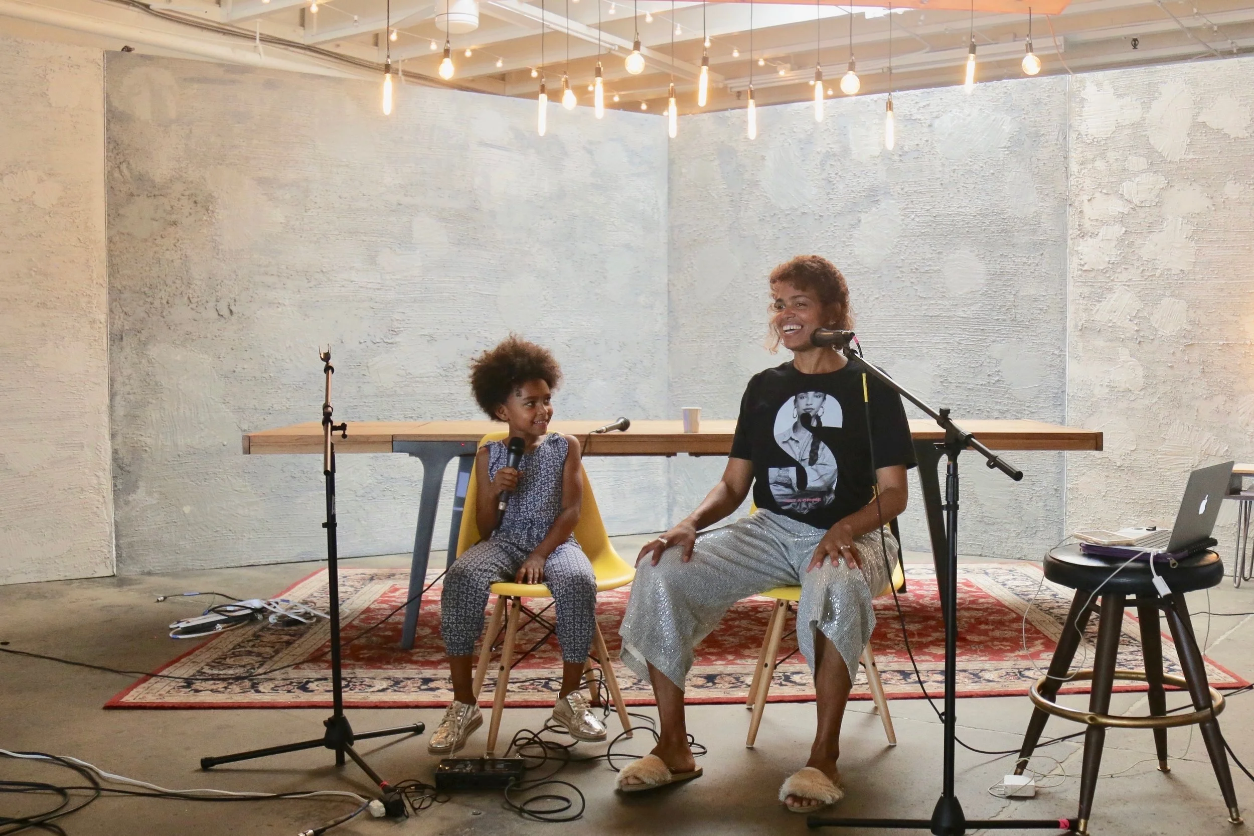 A woman and a young girl sitting on yellow chairs in front of microphones, smiling and talking. The setting appears to be an indoor stage with string lights, a wall with a textured finish, a rug, a table, and audio equipment nearby.