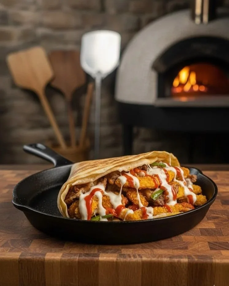 Fried chicken sandwich in a cast iron skillet with toppings, melted cheese, and sauce, with a wood-fired oven and cooking utensils in the background.
