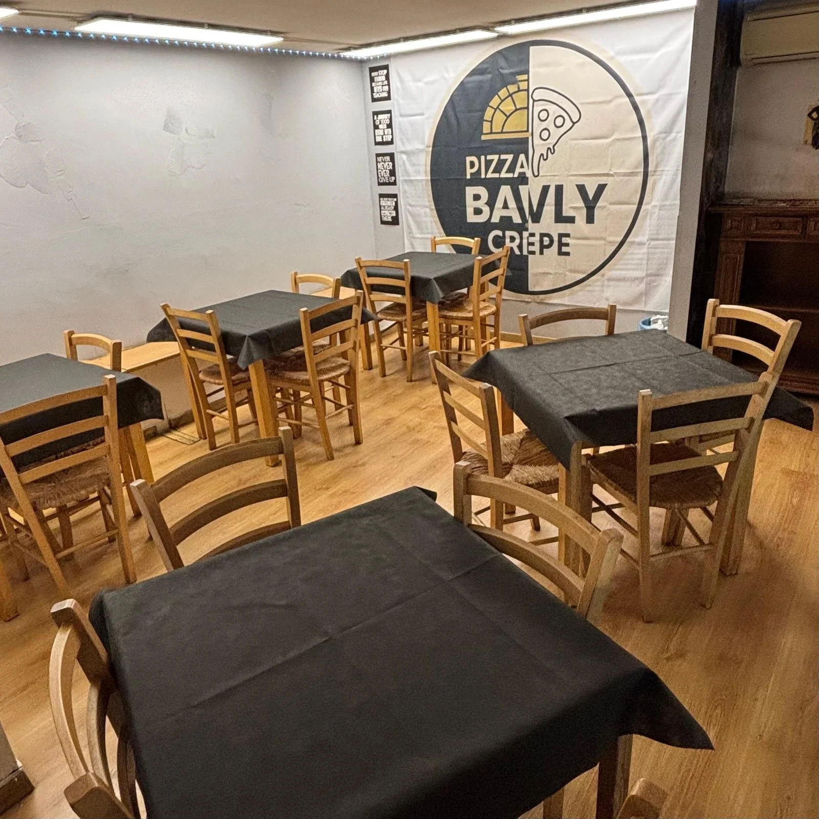 Interior of a pizza restaurant with several tables covered in black tablecloths and wooden chairs. A large mural on the wall displays the restaurant's logo featuring a slice of pizza and the name "Pizza Bawly Crepe."