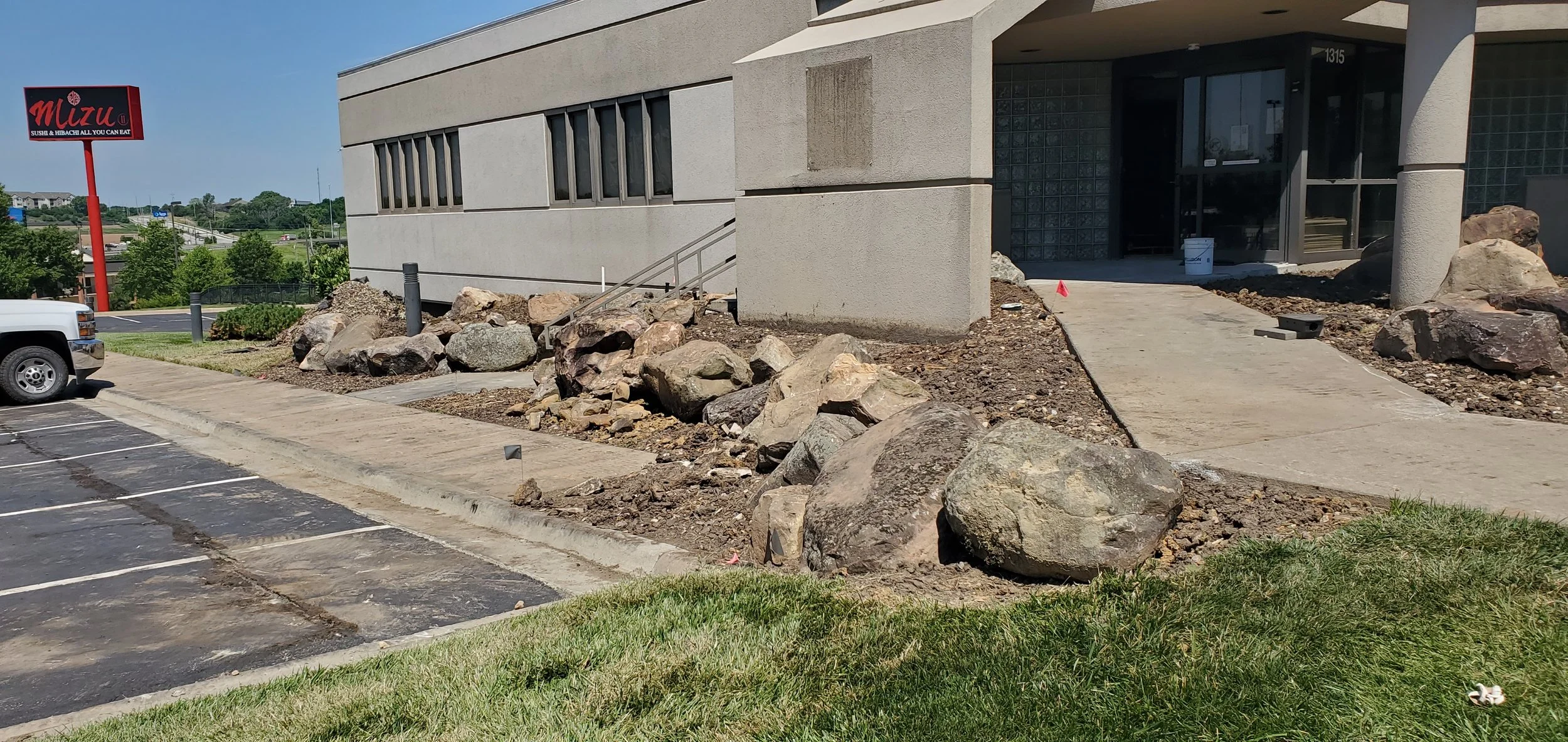Construction area with rocks and dirt outside a commercial building under construction, with a sidewalk and parking lot in front.