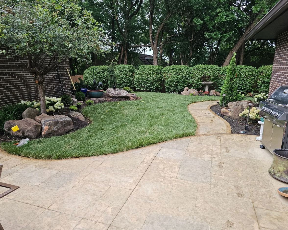 Backyard garden with a curved stone pathway, green grass, trees, bushes, rocks, and a brick house patio with a grill and garden decorations.