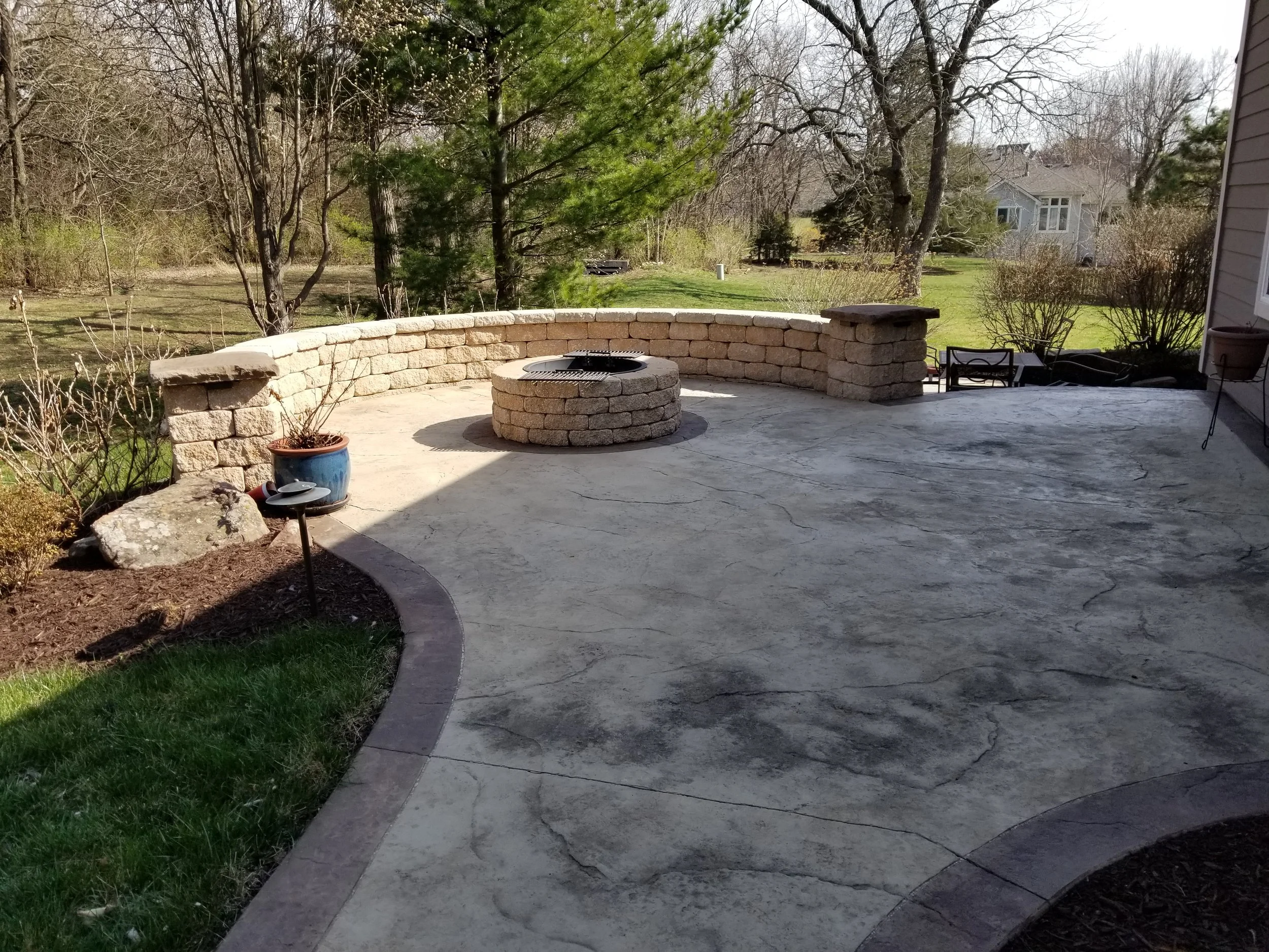 Concrete patio with a curved brick wall, a circular fire pit, and outdoor furniture, surrounded by a backyard with trees and grass.