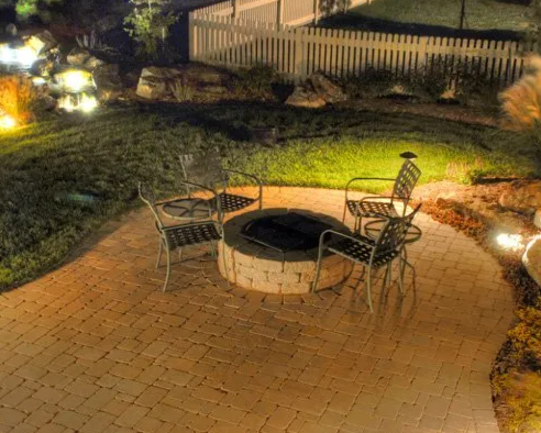 Nighttime outdoor patio with four chairs around a fire pit on a brick surface, surrounded by grass, rocks, and a white fence.