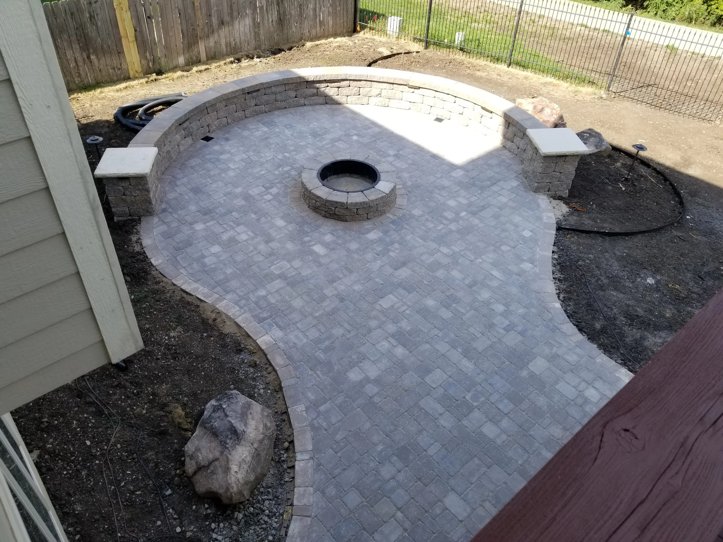 A backyard patio with gray paver stones, a central fire pit, and a curved stone seating wall, surrounded by a dirt area and a black metal fence.