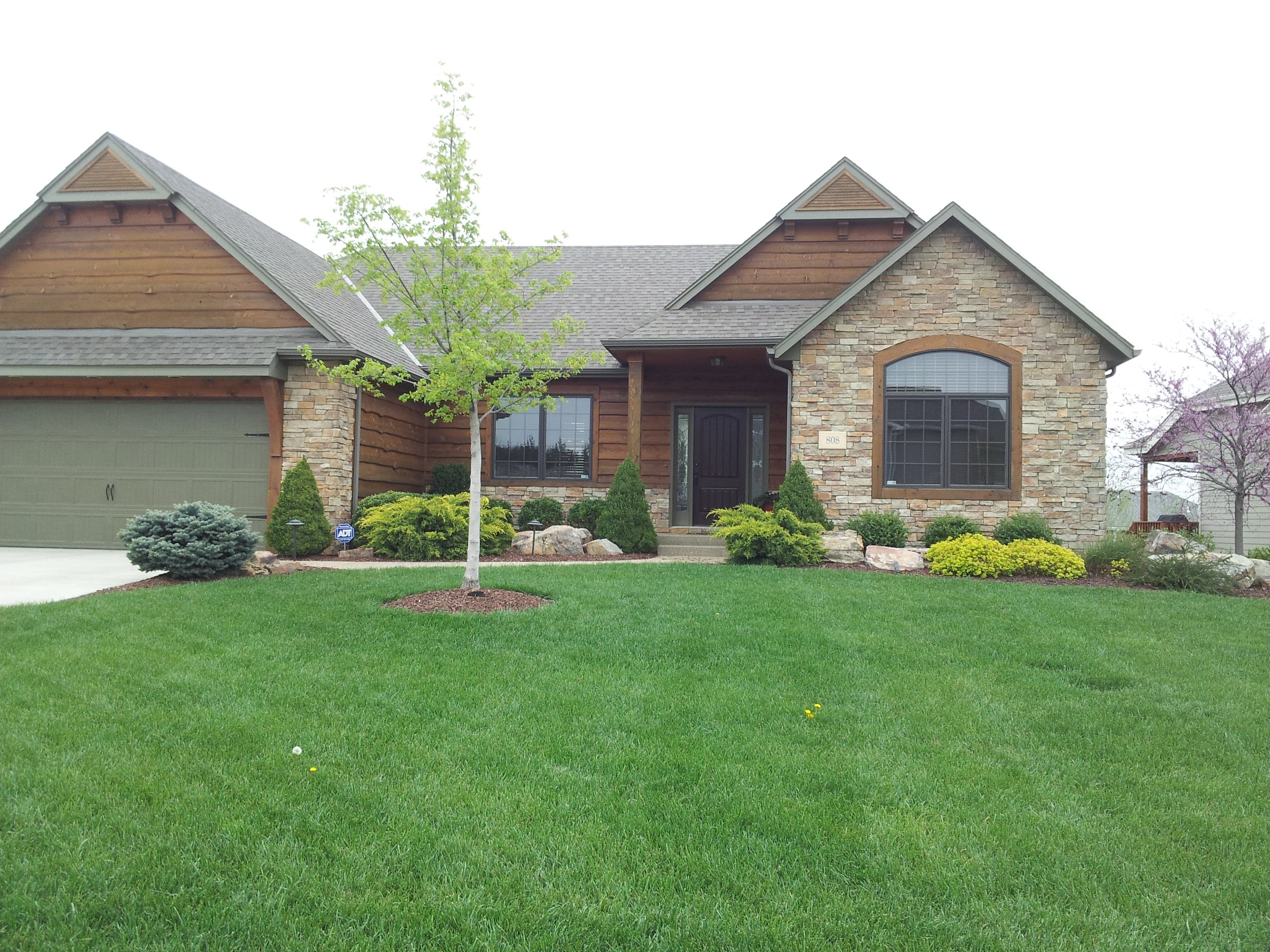 Front view of a house with a well-maintained lawn, small trees, and various shrubs and plants, with a garage on the left side.