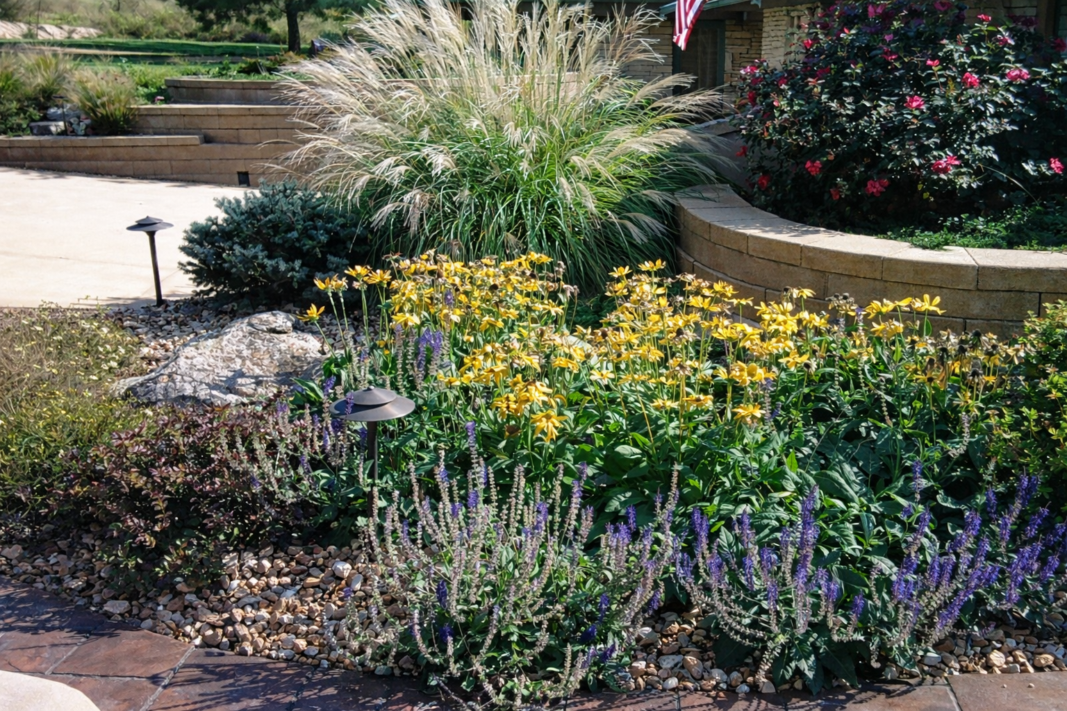 A landscaped garden with yellow, purple, and green plants, small pathway, decorative rocks, brick retaining wall, and garden lights