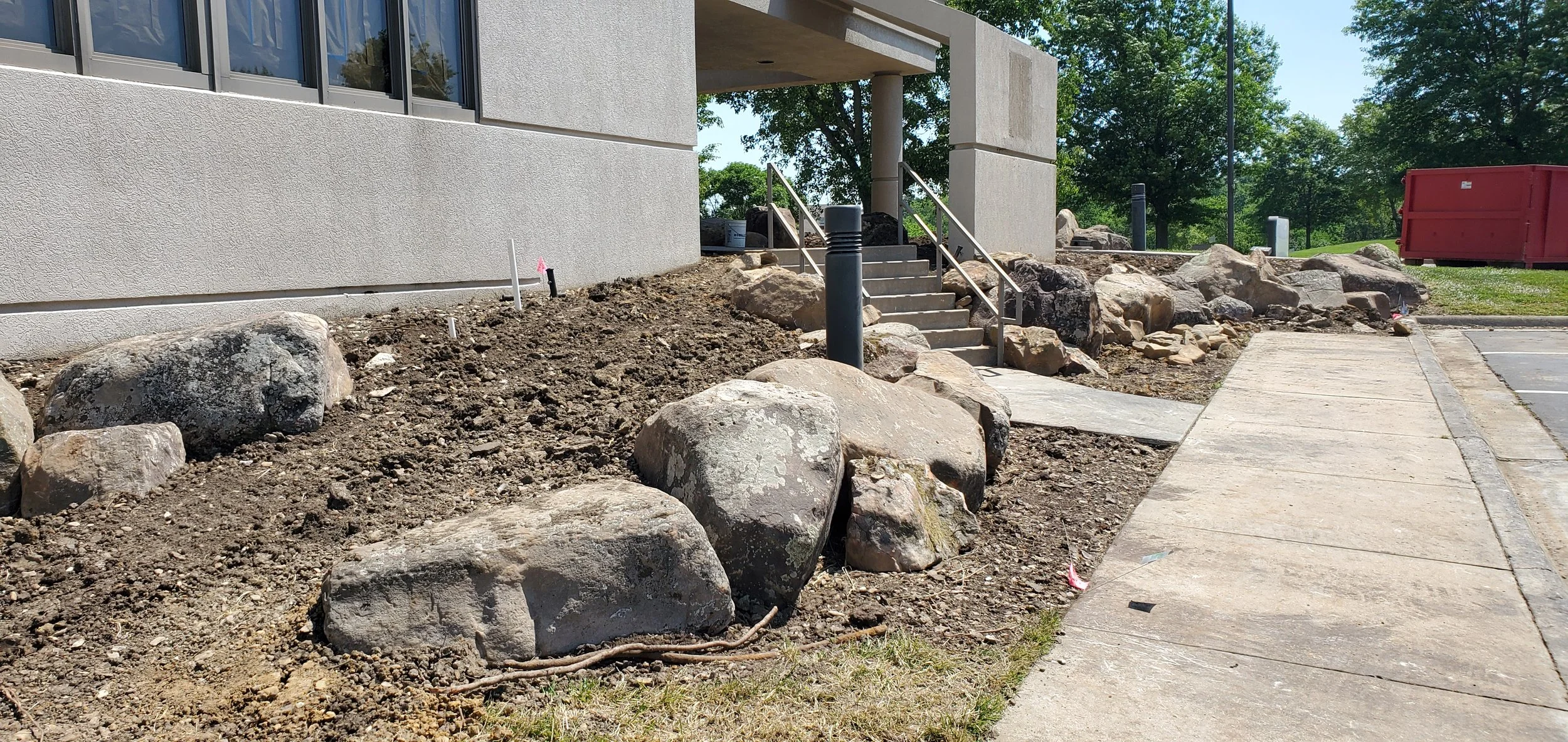 Construction area with large rocks and dirt outside a building, near stairs and a sidewalk, with construction markers and equipment in the background.