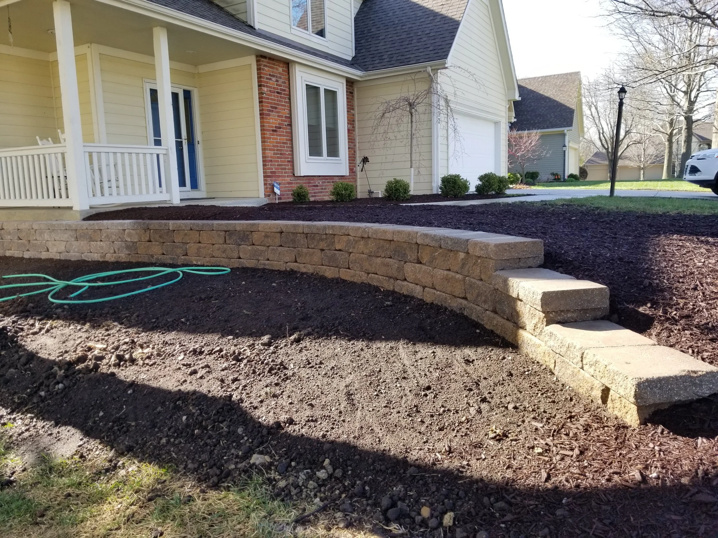 Newly built brick retaining wall and stairs in front of a yellow house with a porch and a lawn in a suburban neighborhood.
