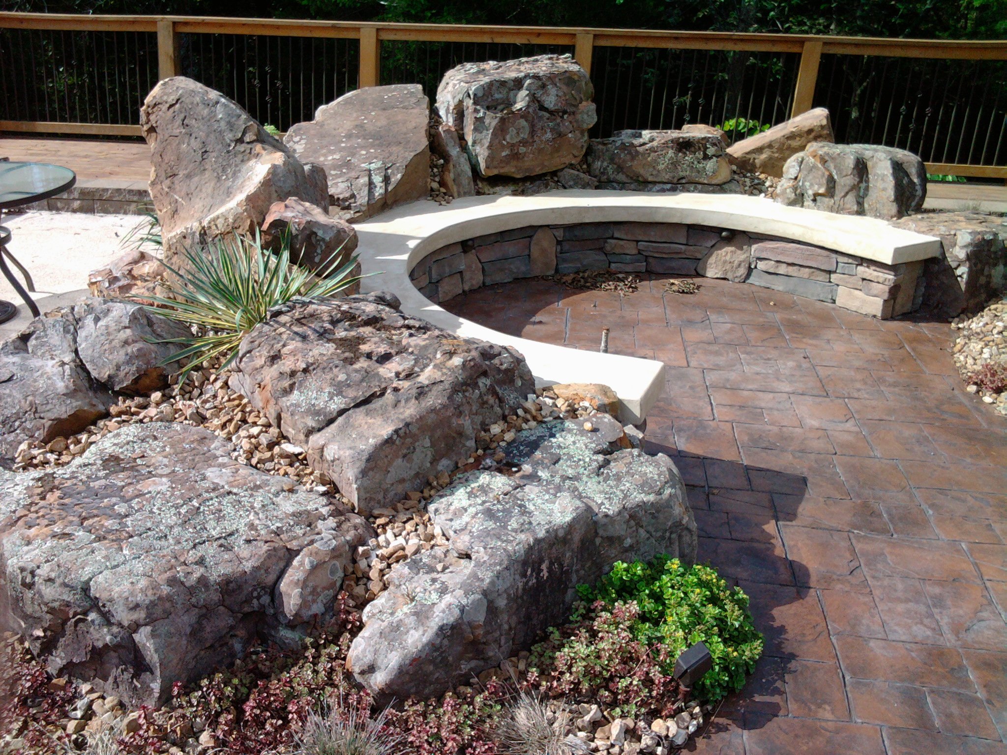 A backyard patio area featuring large rocks, a circular fire pit with a stone wall, a paved surface, and various plants. A wooden railing surrounds the patio.