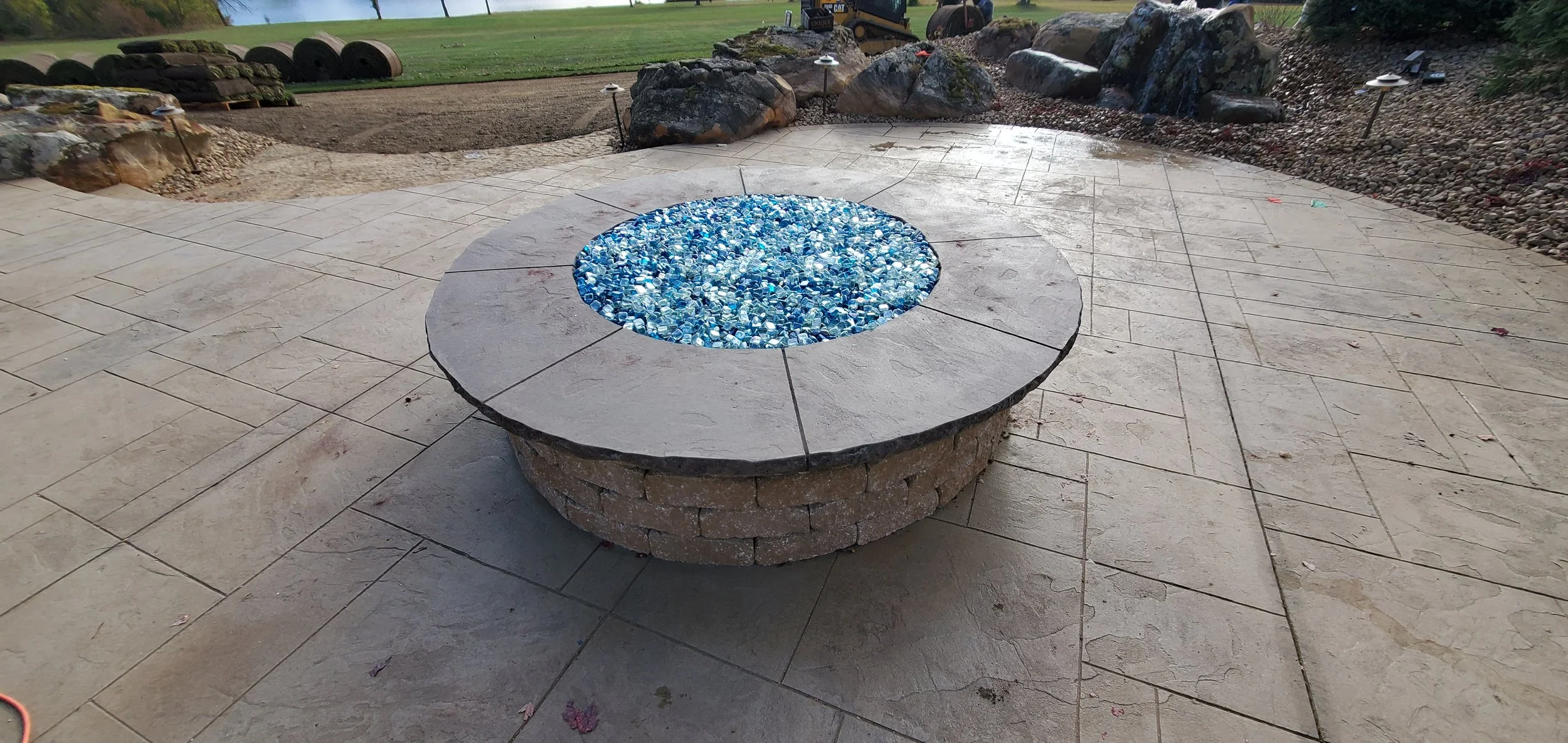 A circular outdoor fire pit with a top made of grey stone, filled with blue glass pebbles. The fire pit has a brick base and is set on a large paved area with additional landscaping and rocks in the background.