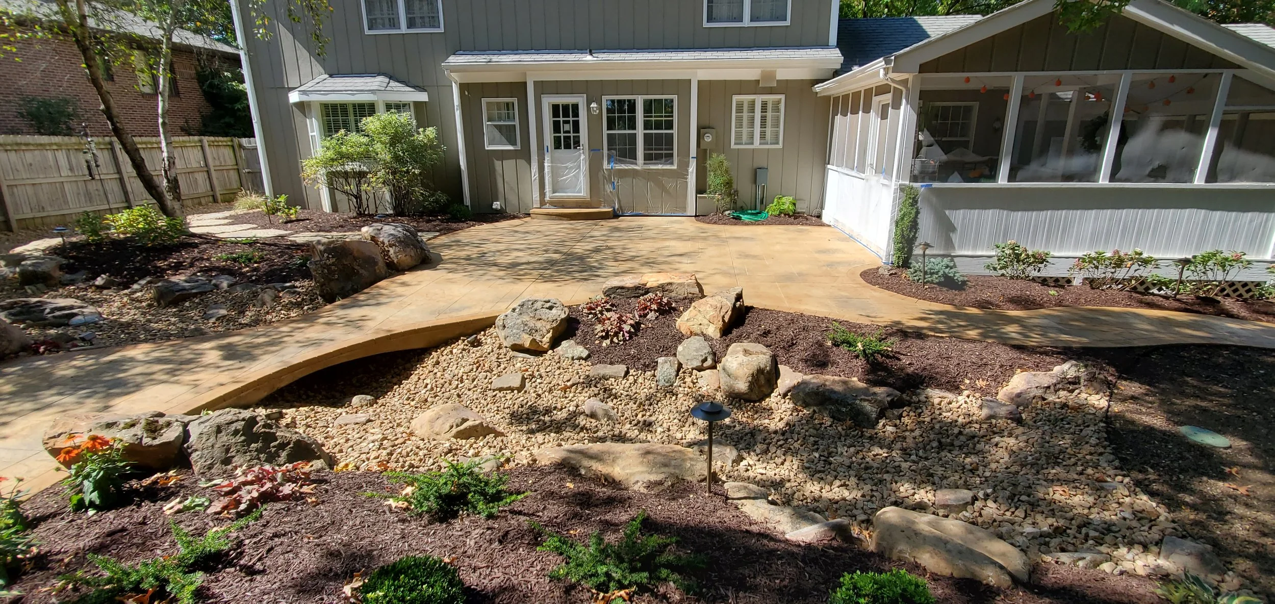 Backyard with a newly paved patio, landscaped garden beds, decorative rocks, and a screened porch attached to a house.