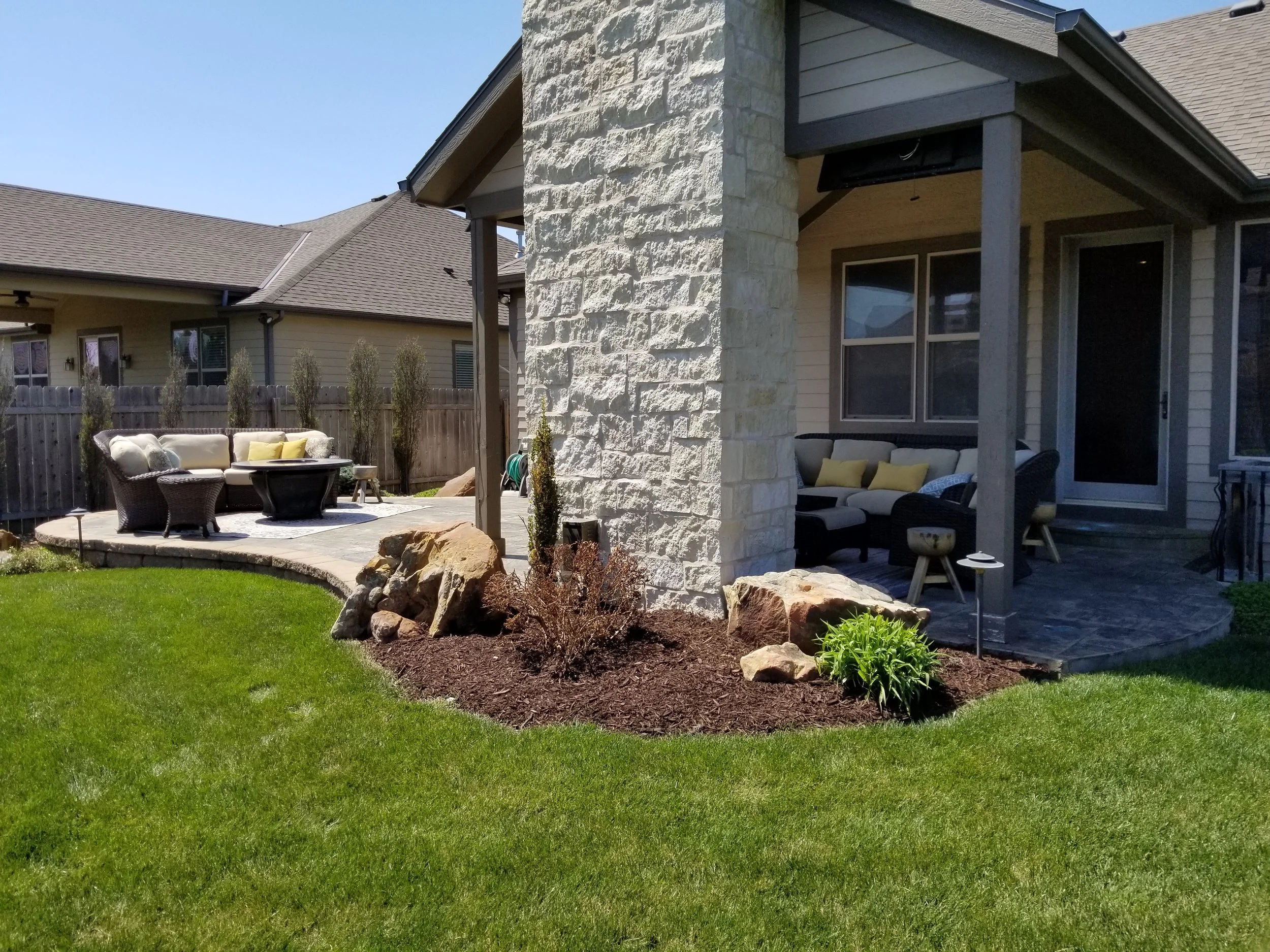 Backyard outdoor patio with seating area, green grass, decorative rocks, and plants, adjacent to a house with stone and siding exterior