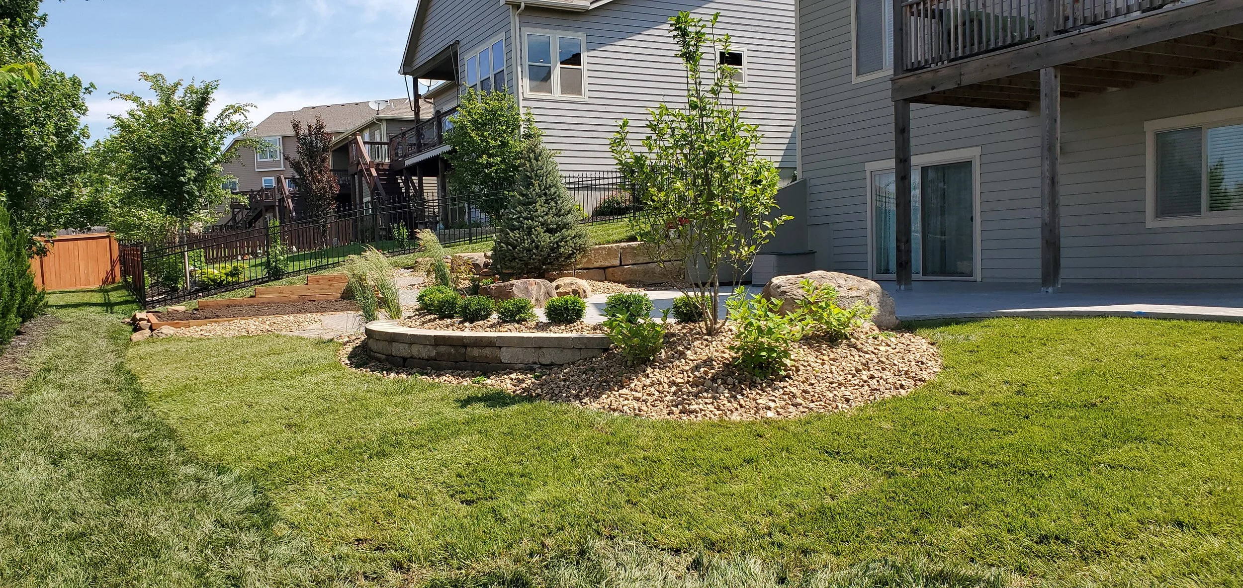 A well-maintained backyard with landscaped garden beds, green grass, small trees, shrubs, rocks, and a patio area attached to a house with sliding glass doors.