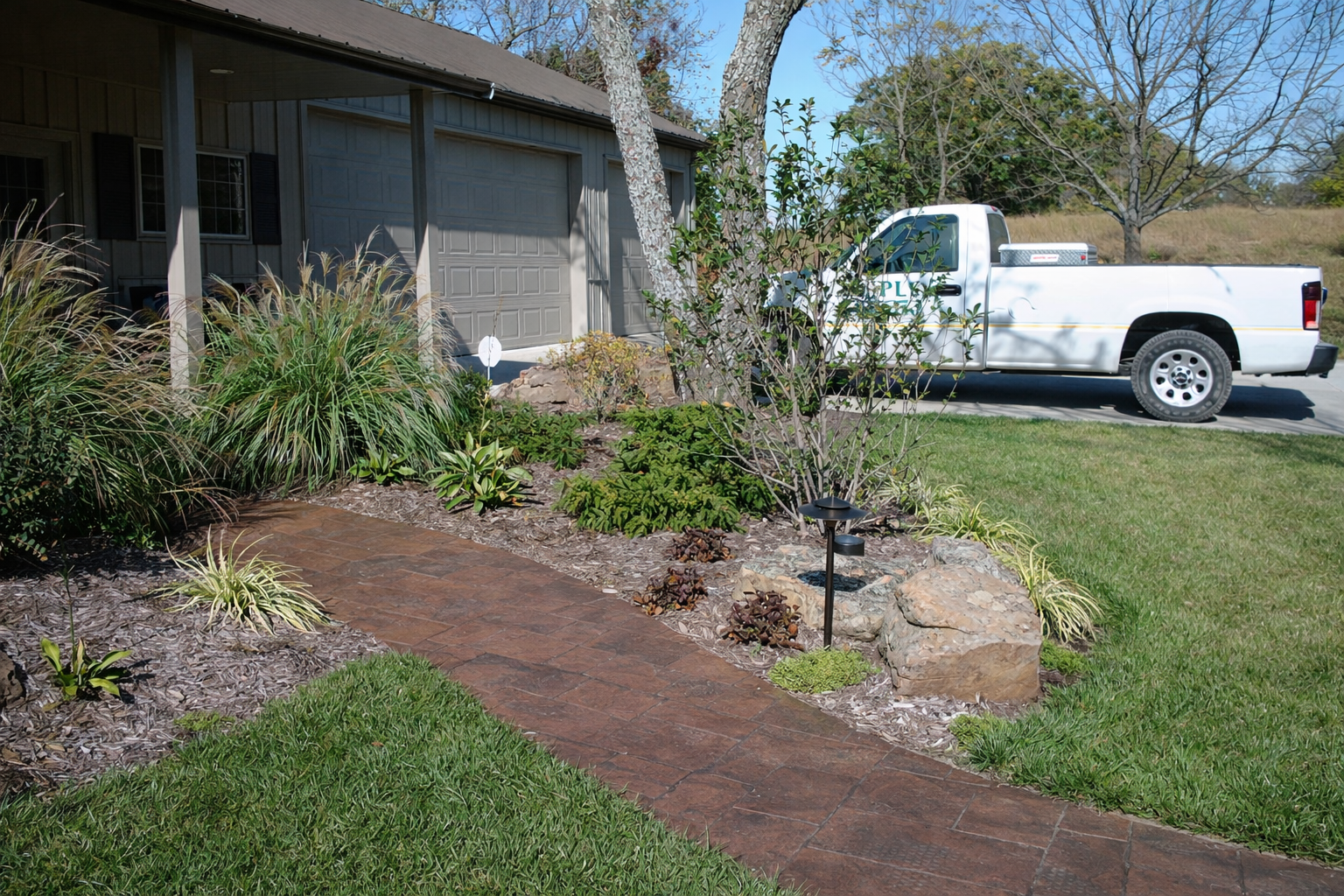 Front yard with a brick pathway, garden bed with shrubs and rocks, and a white pickup truck parked in driveway in front of house with gray garage door and trees in the background.