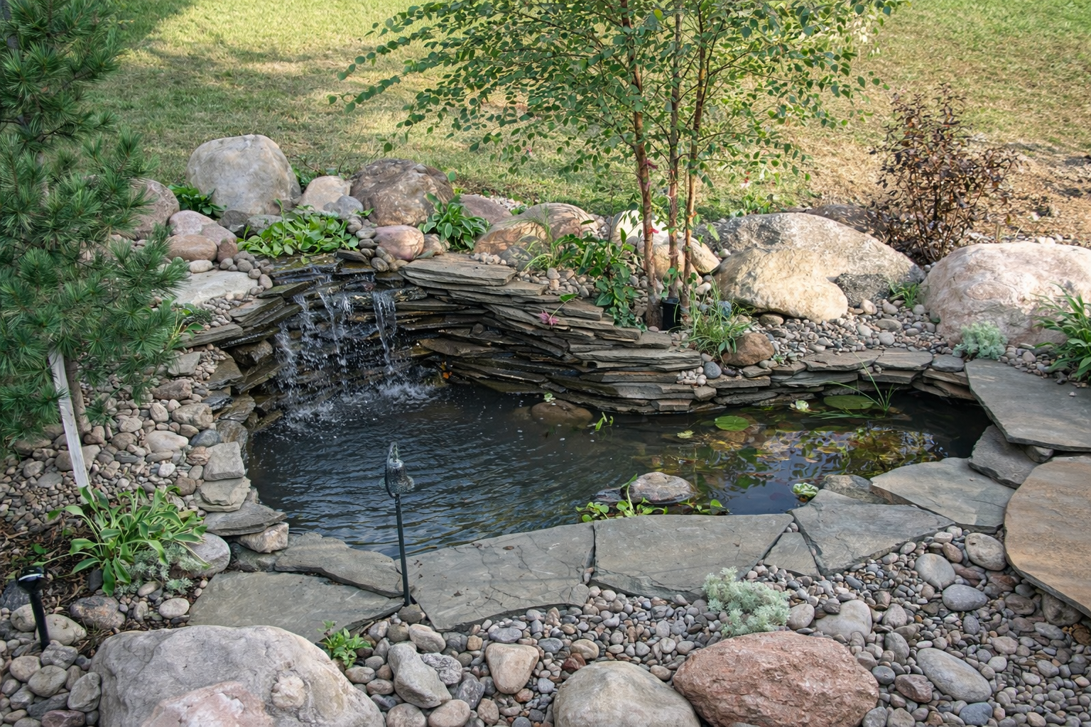Garden pond with small waterfall, surrounded by rocks and plants.