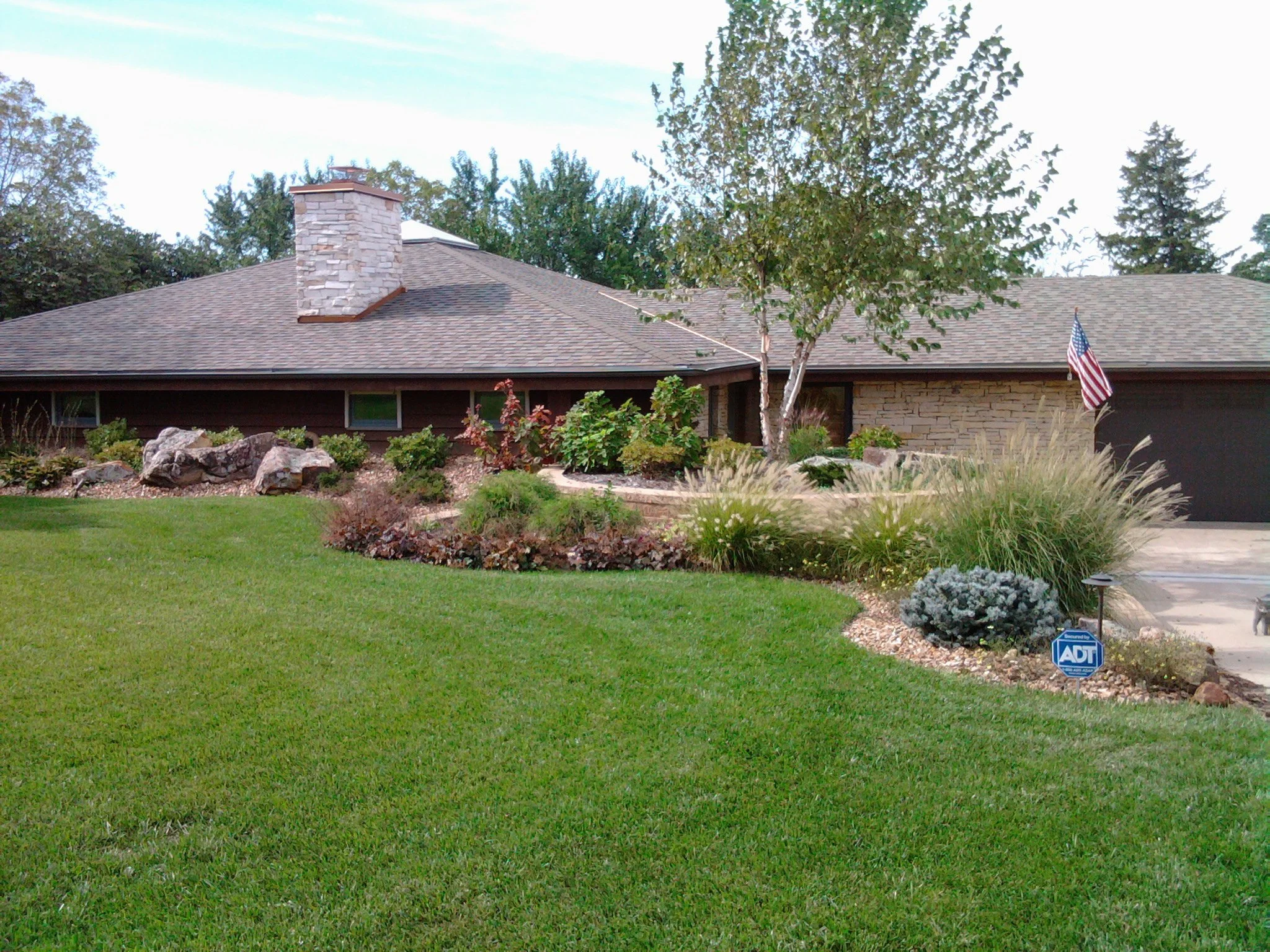 A single-story house with a front yard landscaped with various plants and rocks. There is a tree and an American flag near the driveway. An ADT security sign is visible in the garden bed. The house has small horizontal windows and a chimney, with a b