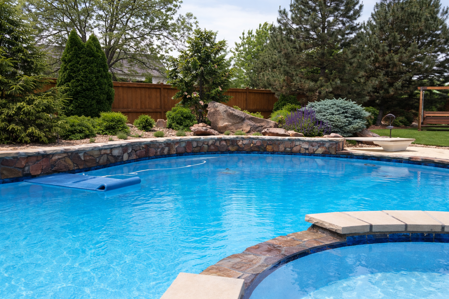A backyard with a swimming pool surrounded by lush green plants, trees, and shrubs, with a brown wooden fence in the background and outdoor seating.