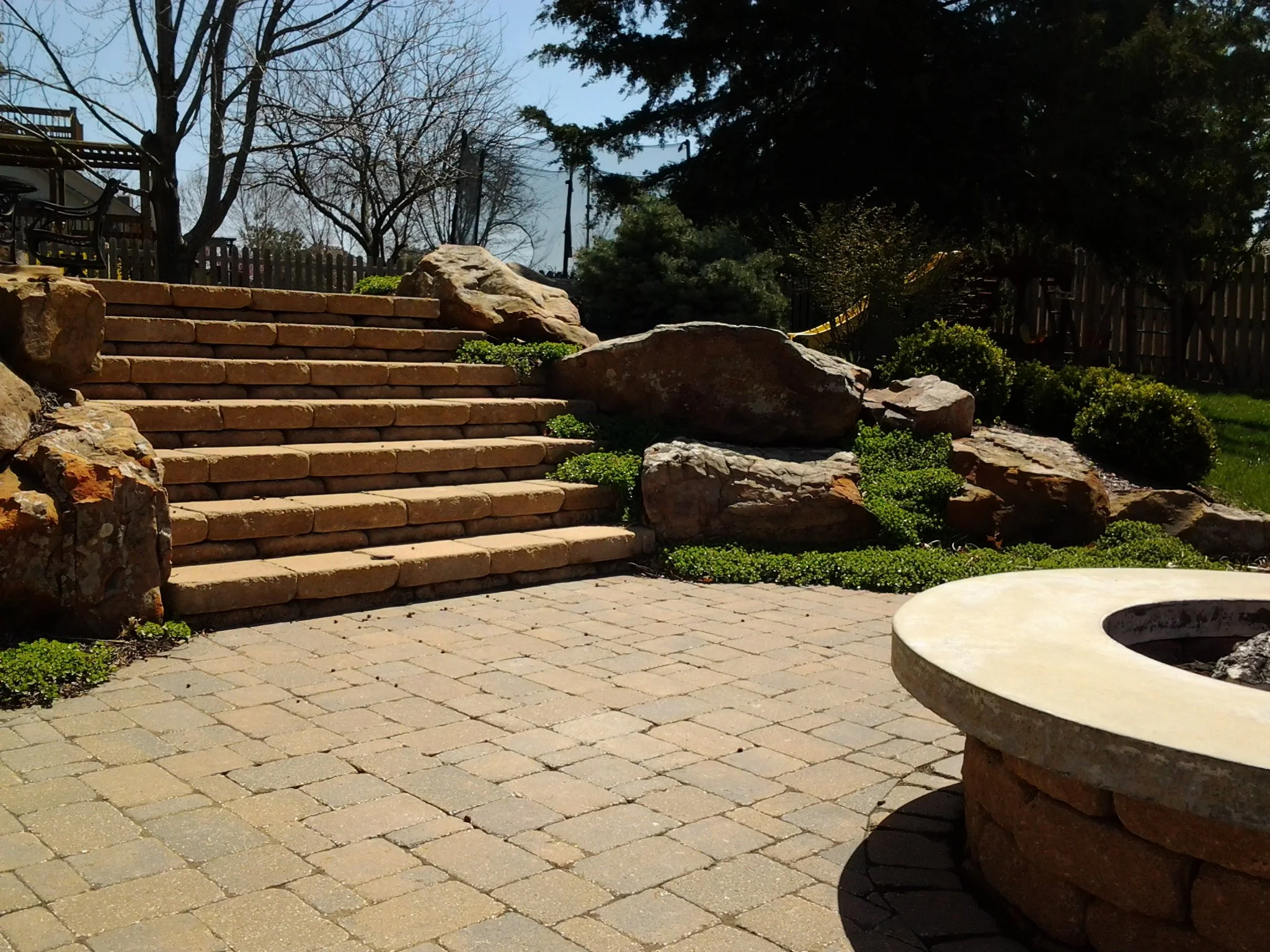 Backyard patio with brick steps leading up to a garden area, surrounded by large rocks and green bushes, with a fire pit in the foreground.