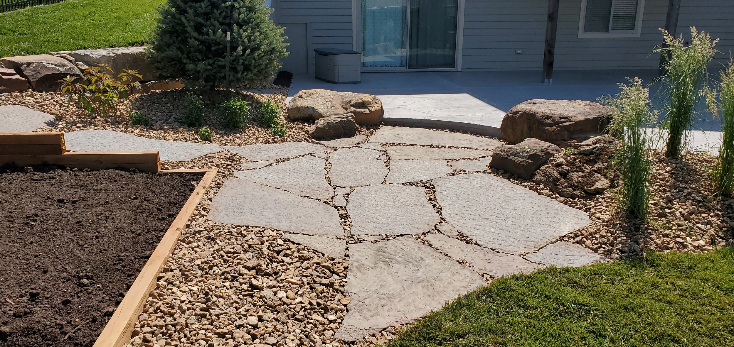 A backyard with a stone pathway leading to a patio, soil in a wooden frame garden bed on the left, and various rocks, plants, and a house in the background.