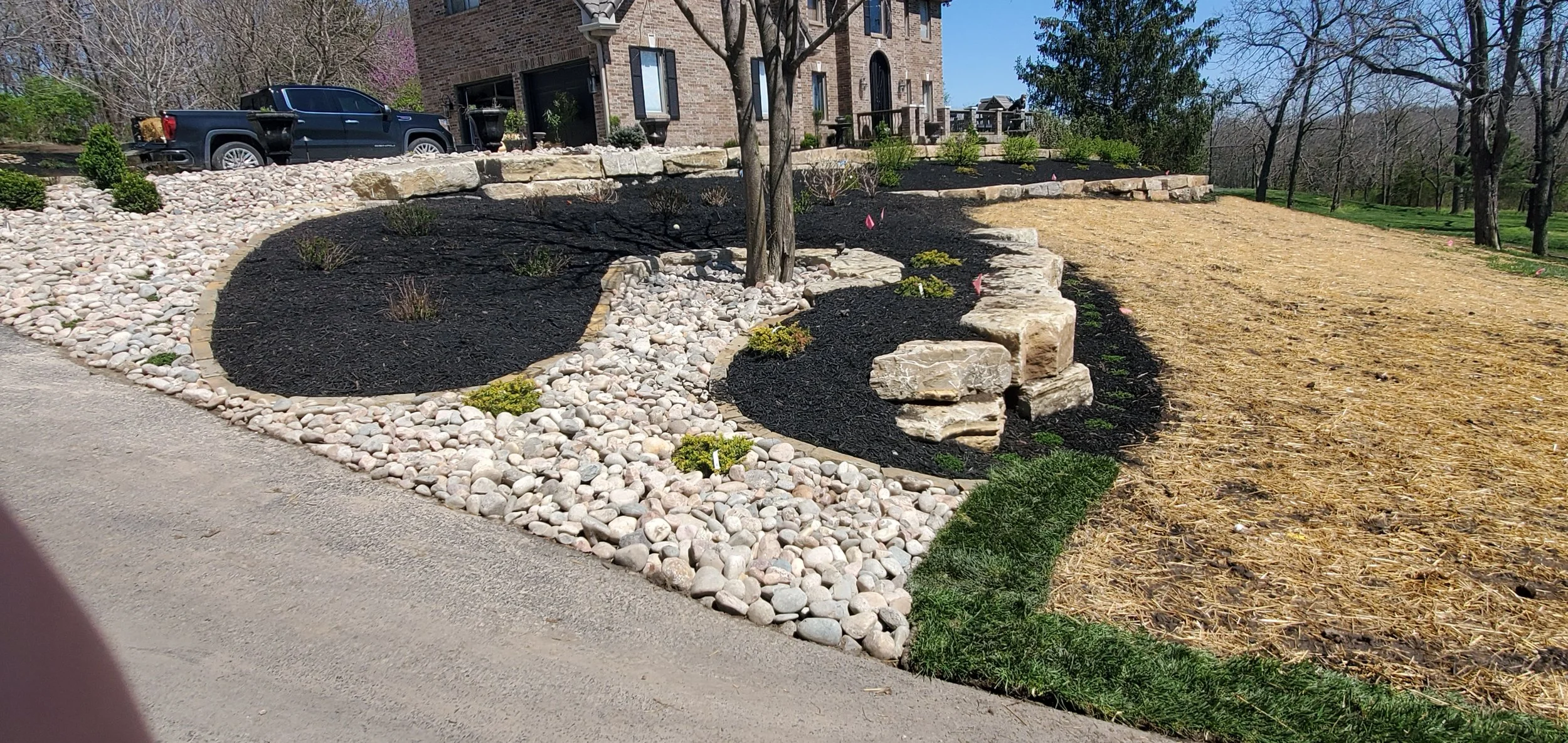 Residential landscaping with layered stone mulch, small plants, and a tree in the yard.