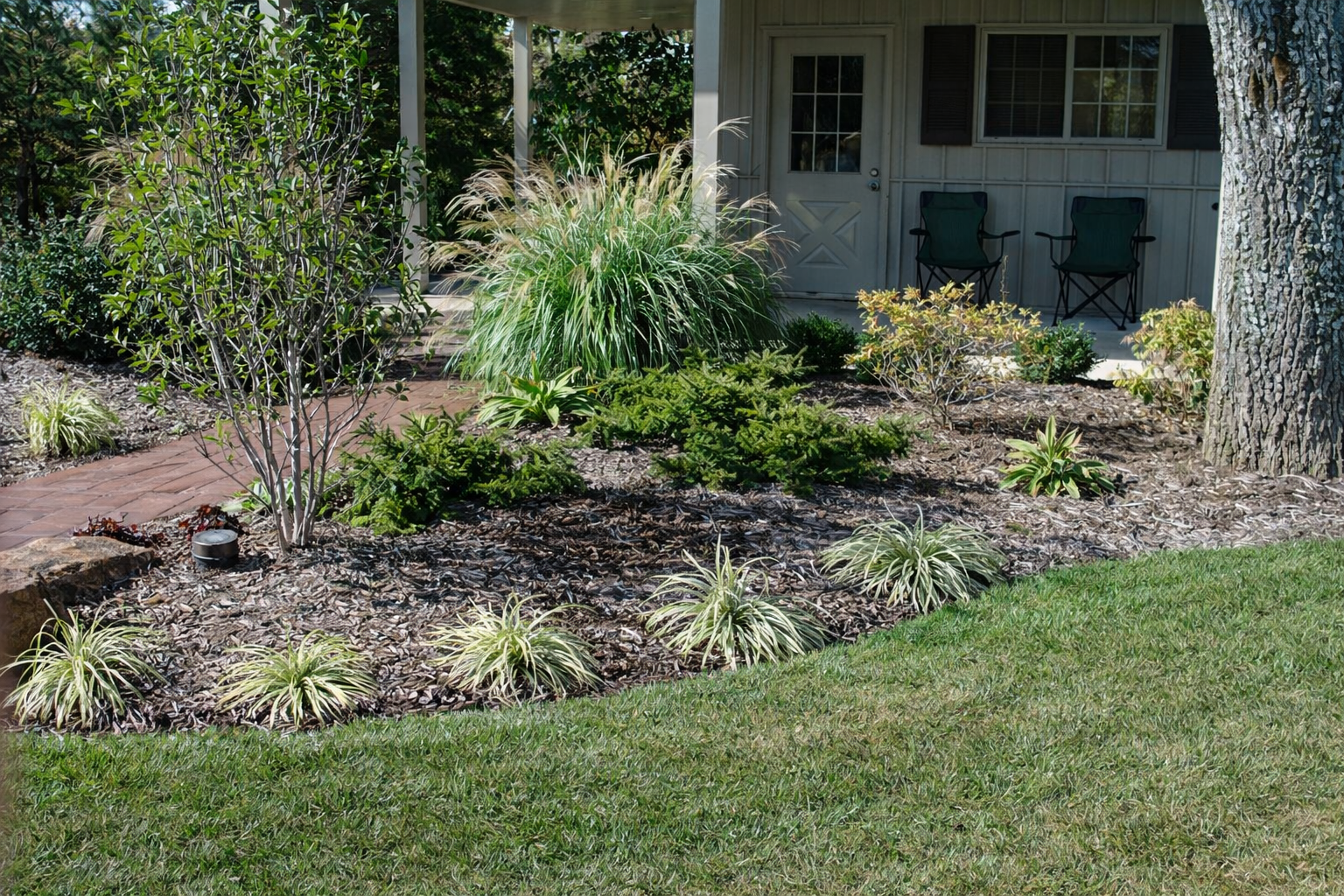 A landscaped front yard with a brick pathway, various green plants and shrubs, mulch covering the garden beds, a grassy lawn, and a porch with chairs and a window in the background.