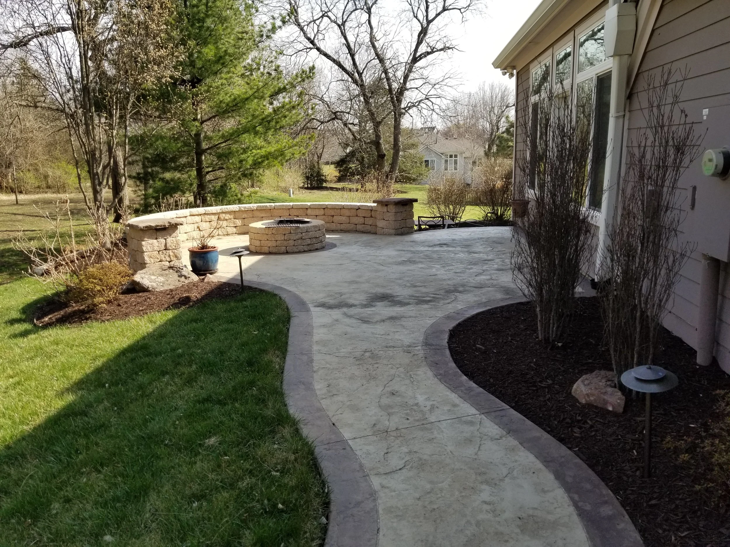 Backyard patio with curved concrete walkway, grassy area, and a stone fire pit. The patio is bordered by mulch beds with shrubs and outdoor lighting, with trees and neighboring houses in the background.