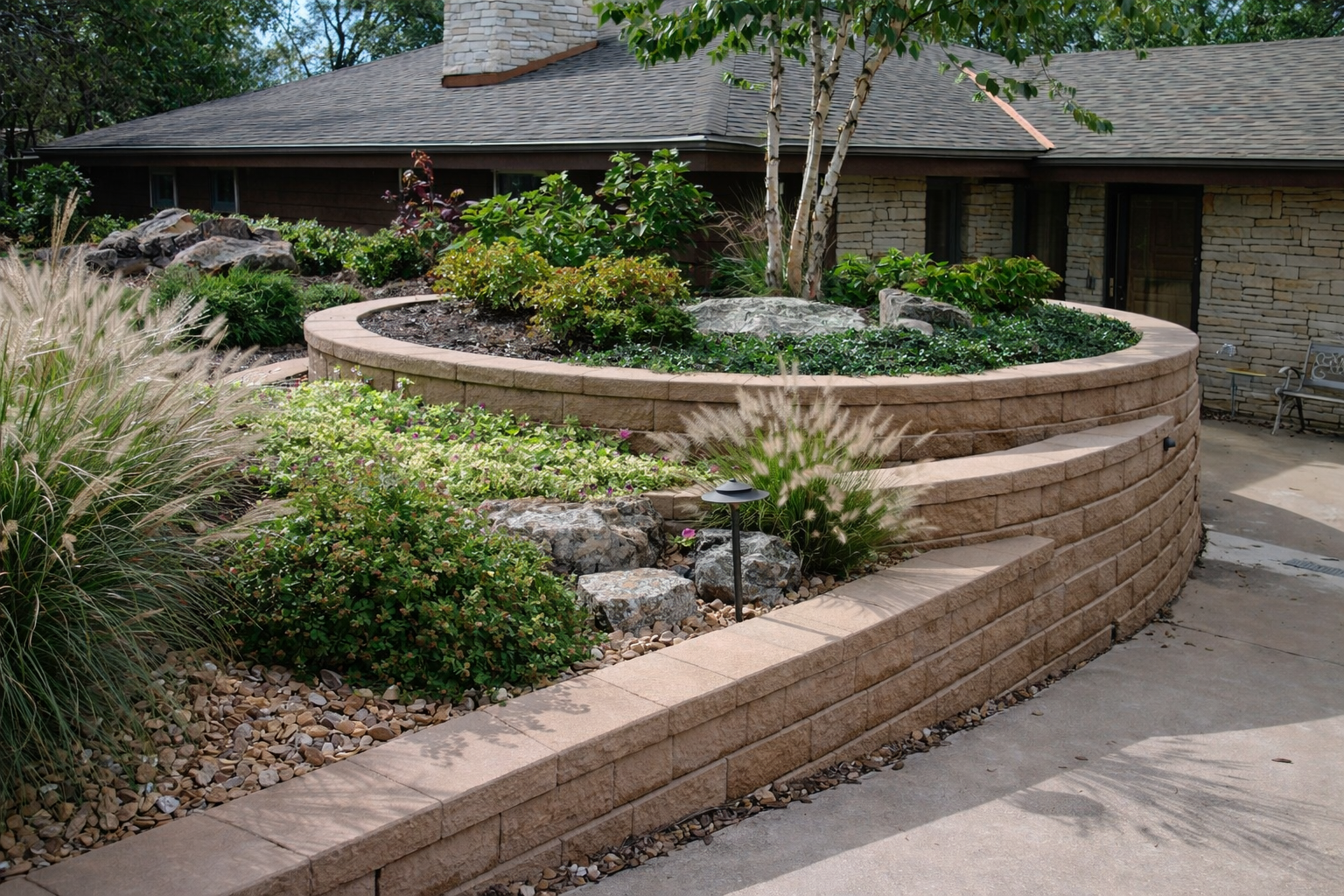 A landscaped backyard with a curved raised flower bed made of beige bricks, filled with various green plants, shrubs, and small trees. There are rocks and gravel around the plants, and a garden light. A patio area and part of a house with stone and w