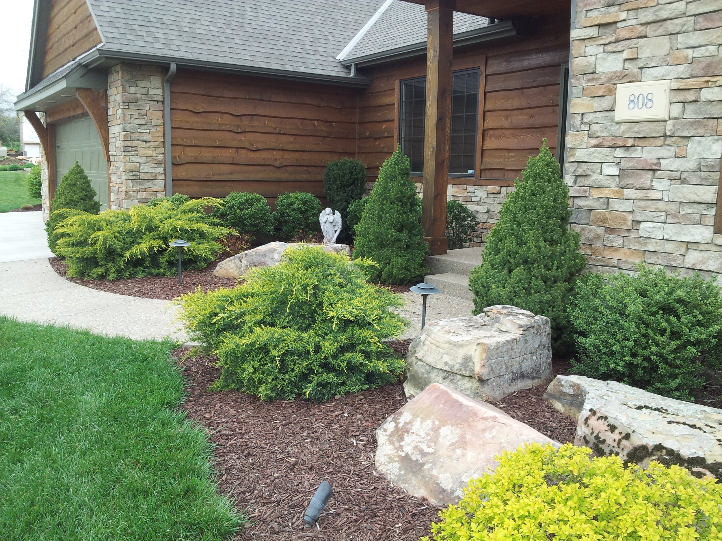 A front yard garden with various green bushes, rocks, and pathway with lights next to a house with a garage. The house has wooden siding, stone accents, and the number 808 on the wall.