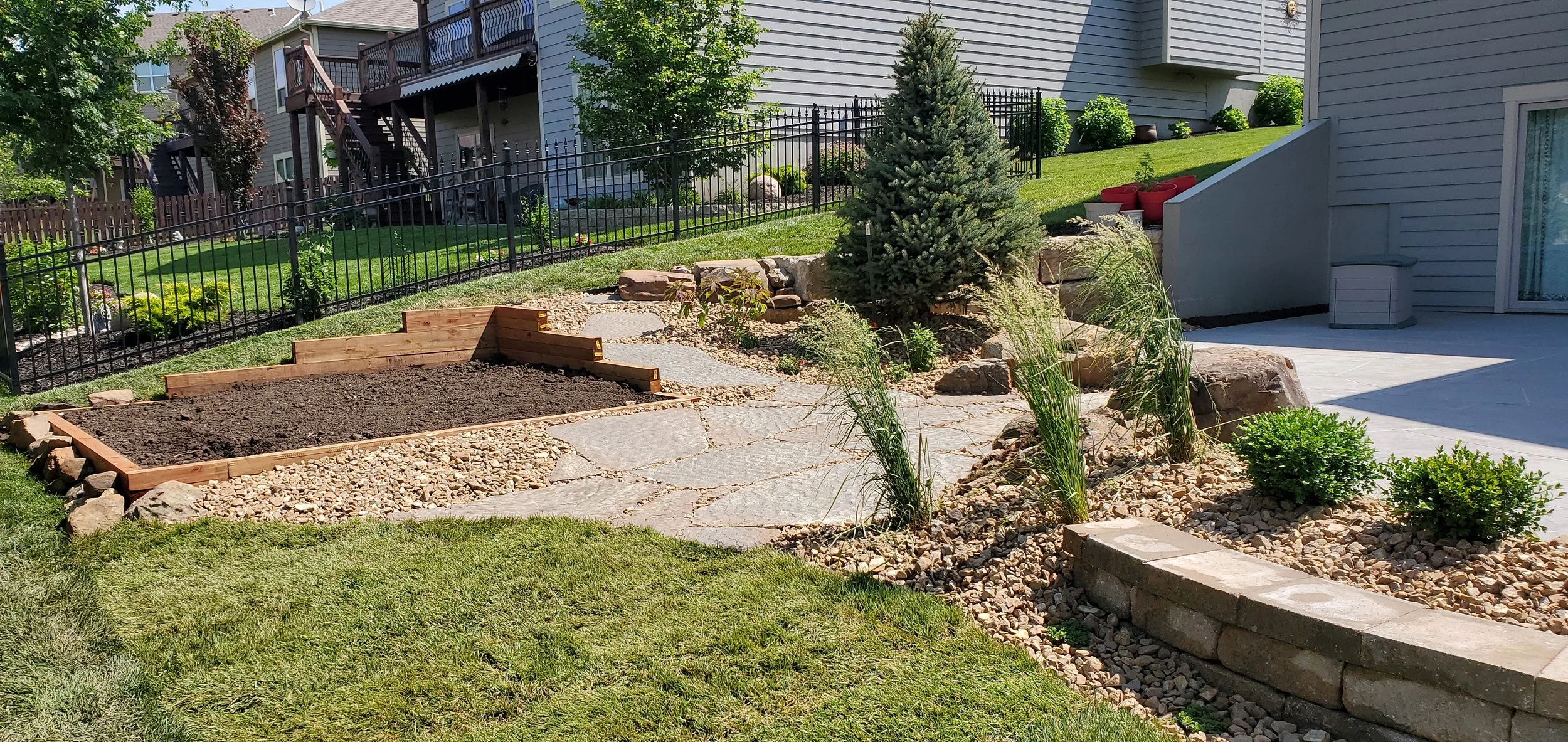 A landscaped backyard with a small garden bed, a stone pathway, a grassy area, and a patio. The garden bed contains plants and rocks with a large evergreen tree nearby. A black metal fence surrounds the yard, with adjacent houses and trees.
